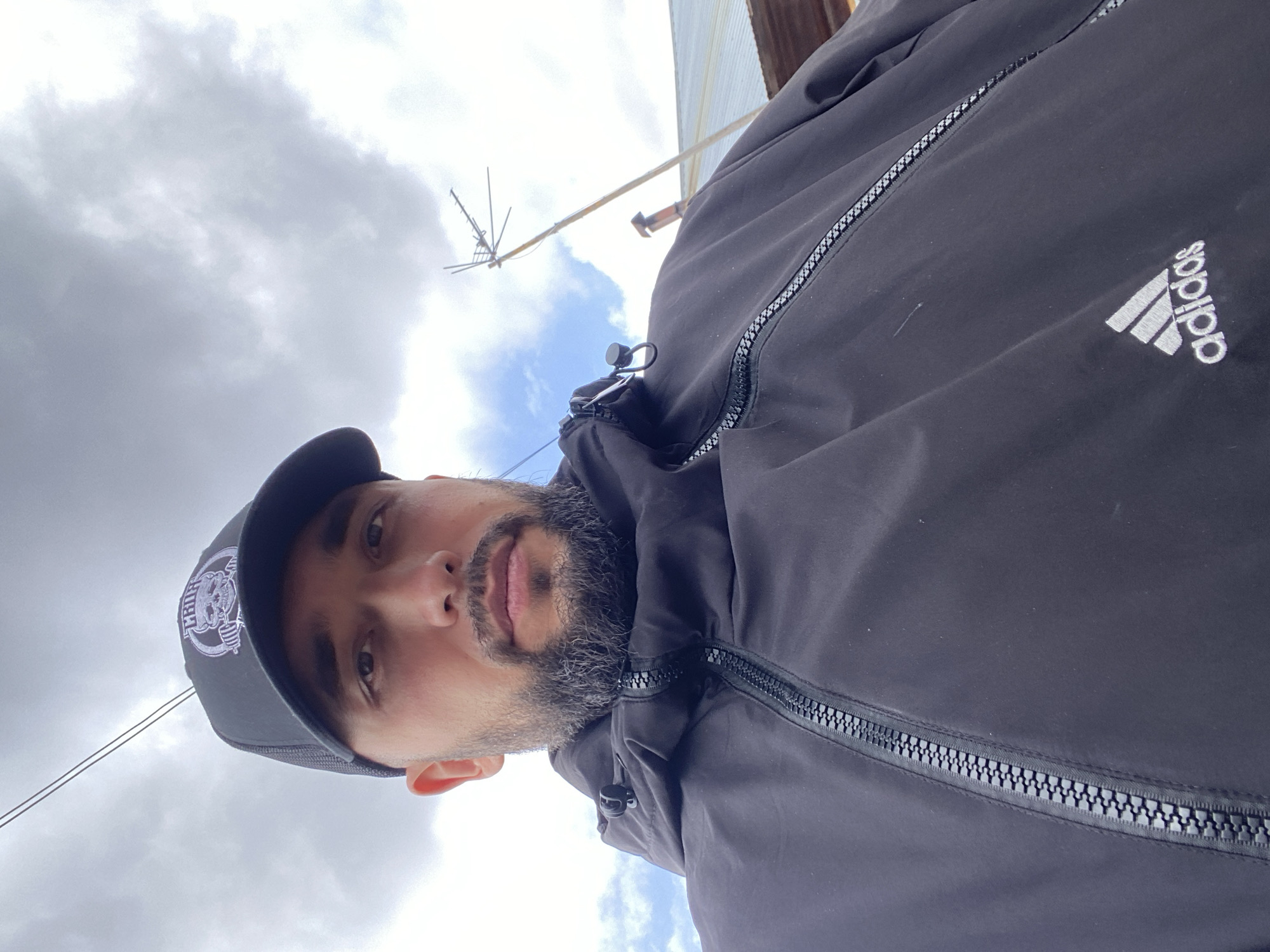 A man stands outside, wearing a black Adidas jacket and a dark baseball cap with a logo. He appears to be looking down at the camera, suggesting the photo was taken from a slightly higher angle. The background shows a cloudy sky, a utility pole and a building. There is insufficient information to verify if the location is Zhigalki, Russia.