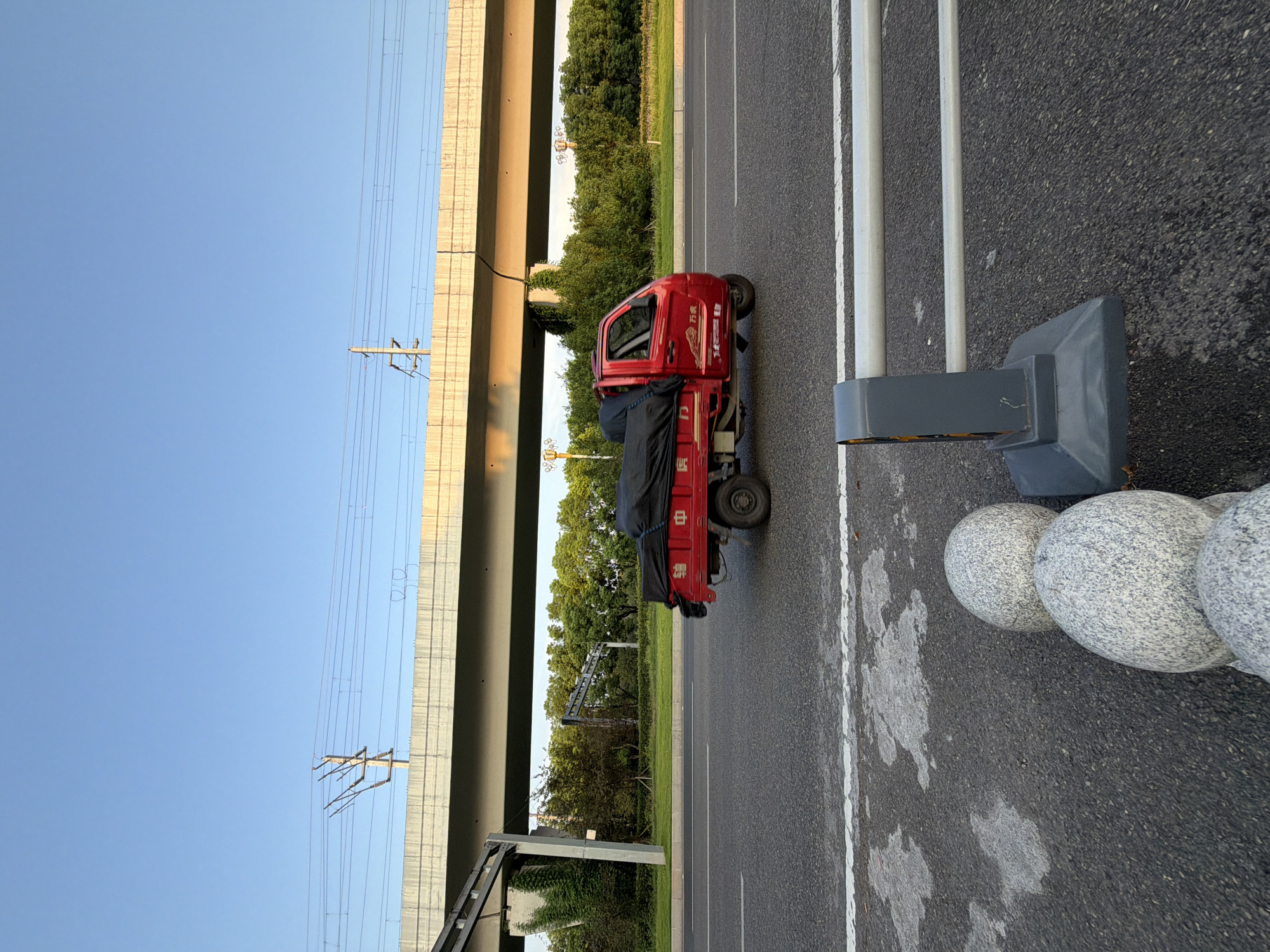 A red three-wheeled truck is driving on a road under an elevated railway bridge. The truck is covered with a black tarp. The sky is clear and blue, and trees line the side of the road. In the foreground, there are three round, granite bollards and a grey barrier. The image appears to be taken in the city of Jiangjin, China.