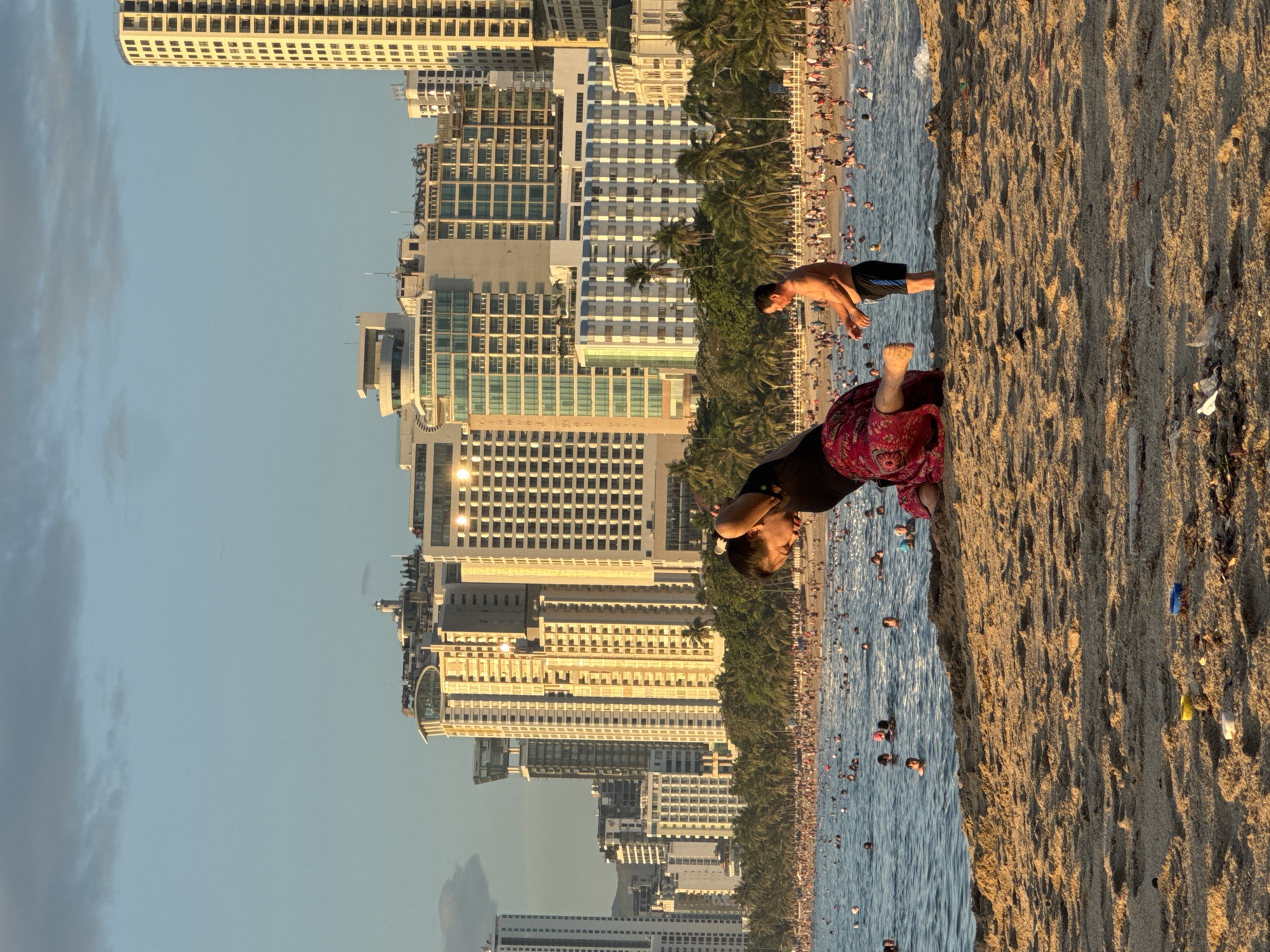 This is a wide-angle, eye-level, outdoor, daytime photograph taken on a sandy beach in Nha Trang, Vietnam. In the foreground, a person is seated on the sand, facing away from the viewer, with their head bent forward as if performing a yoga pose or stretching. They are wearing a black top and a patterned red skirt. To the right of the seated person, a shirtless man stands facing the water, wearing black swim trunks with a blue stripe.
The middle ground features a crowded beach with numerous people swimming and sunbathing in the ocean. The water is blue and appears to have moderate waves. A line of palm trees and lush green foliage separates the beach from a backdrop of tall, modern buildings.
The background is dominated by a dense urban skyline with numerous high-rise buildings. The buildings vary in design and color, with many appearing to be hotels or apartment complexes. The sky above is a clear blue with some wispy clouds. The overall impression is a bustling coastal city scene during a sunny day.