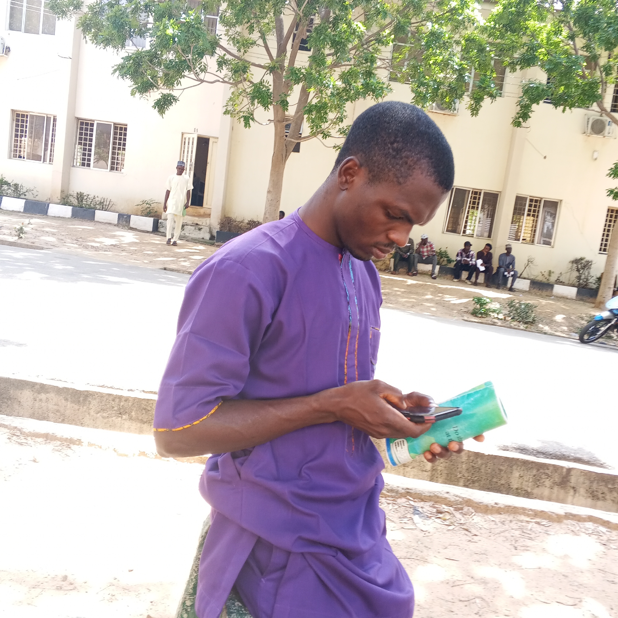 A young man wearing a purple traditional outfit stands outdoors in front of a light-colored building, possibly a school or office. He is holding a book or notebook in his left hand and a mobile phone in his right hand, looking down at them. The building has several windows and is partially shaded by a leafy tree. In the background, other people are seated on a low wall or bench. A motorcycle is parked to the right of the frame. The ground is paved with concrete and dirt. The scene appears to be in Arawa, Nigeria.