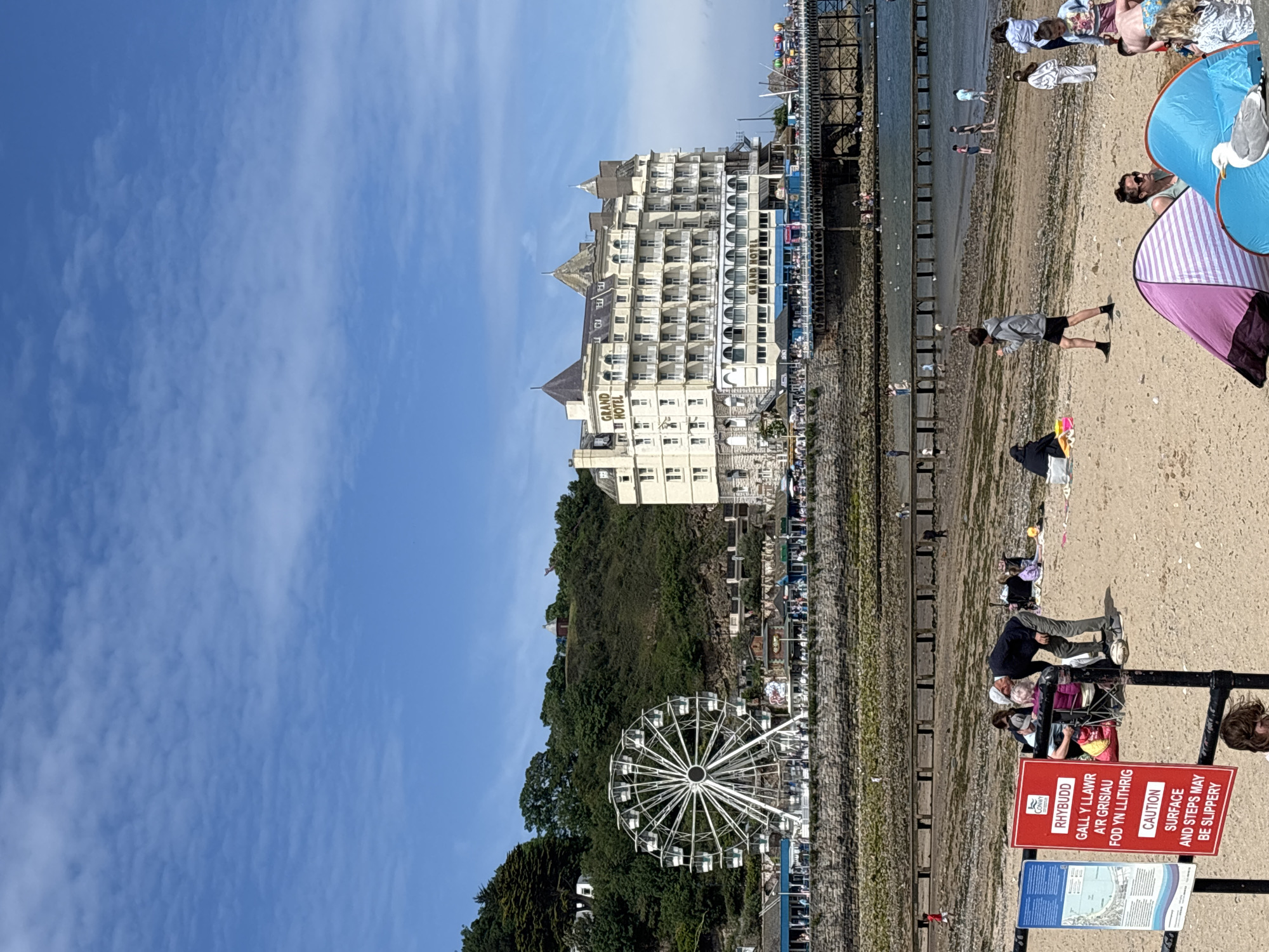 The image shows a seaside scene in Llandudno, United Kingdom. The Grand Hotel dominates the view, its multi-storied facade rising behind a tree-covered hill where a Ferris wheel is located. A pier extends into the sea, with people visible along its length. A beach is present, with people relaxing, walking, and engaging in activities. A caution sign warns of slippery surfaces. The sky is blue with scattered clouds.