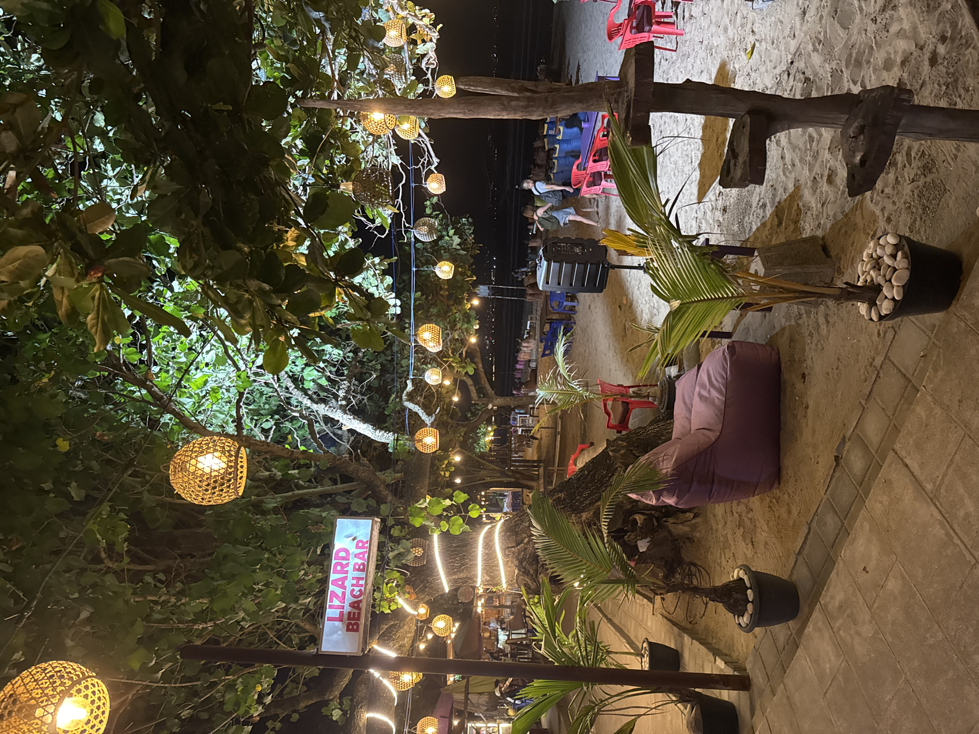 The image depicts a beachside bar named "LIZARD BEACH BAR" in Kuta, Indonesia, at night. The foreground features a sandy area with potted palm trees and a large purple beanbag chair. To the left, a tiled walkway lines the sand. A wooden pole with tiered platforms stands on the right. The background reveals the beach with numerous tables and chairs, some with red seats. Several people are visible, including two women walking towards the water and other patrons seated at tables. Woven lamps hang from trees and wires, casting a warm glow, and string lights are wrapped around a large tree trunk. The sea is visible in the distance under a dark sky.
