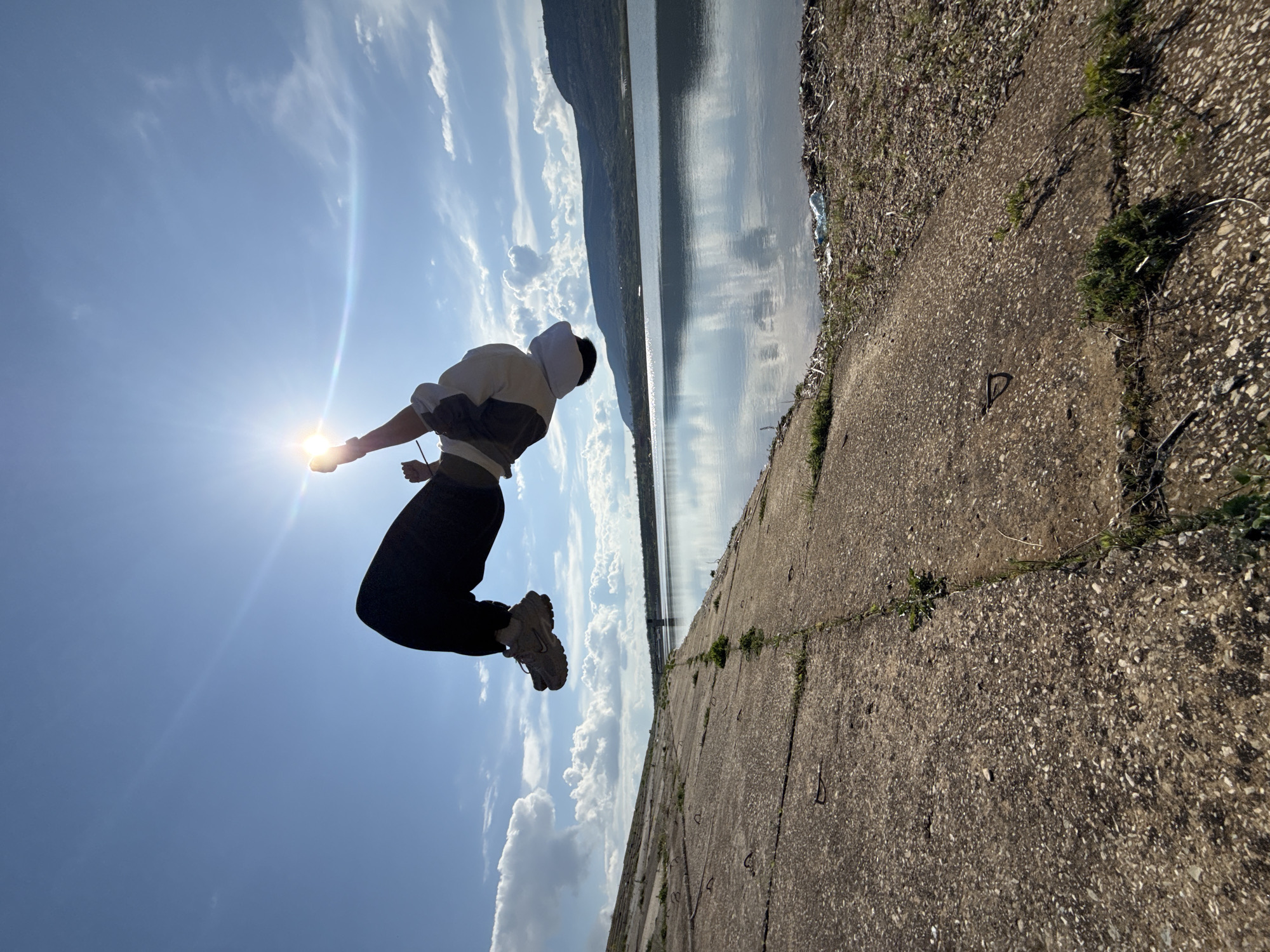 The image captures a person mid-air against a bright, sunlit sky with scattered clouds. The person is wearing a light-colored hooded jacket with darker sleeves, dark pants, and athletic shoes. The person appears to be jumping or in motion with one arm outstretched towards the sun.
The background features a body of water reflecting the sky, likely a lake or reservoir, with distant hills or mountains visible across the water. A concrete embankment or path with cracks and some vegetation runs along the right edge of the frame, suggesting a man-made structure near the water.
The location appears to be near a body of water, potentially a reservoir or lake, with a concrete embankment. It is identified as being located in or near the city of Shumen, Bulgaria.