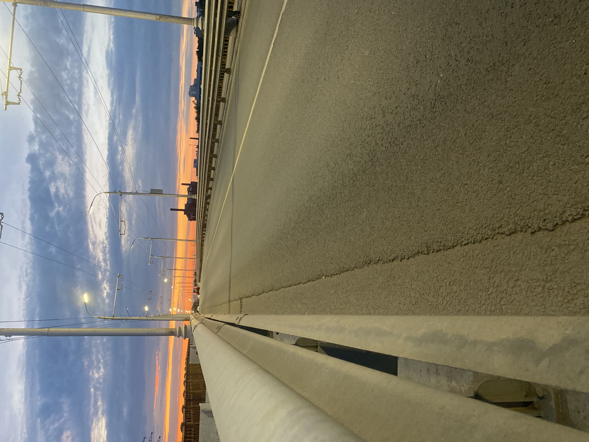 The image depicts a street-level view of a bridge at sunset in Novokuybyshevsk, Russia. The sky is painted with hues of orange and blue from the setting sun, with dramatic clouds overhead. Streetlights line the bridge, casting a soft glow on the asphalt road, which has a white line marking its center. A metal guardrail runs along the side of the bridge, separating the road from a concrete barrier. In the distance, industrial buildings with smokestacks are visible, along with a cluster of residential buildings. A single car is driving away from the camera on the bridge. The overall scene suggests a transitional period between day and night in an urban industrial setting.