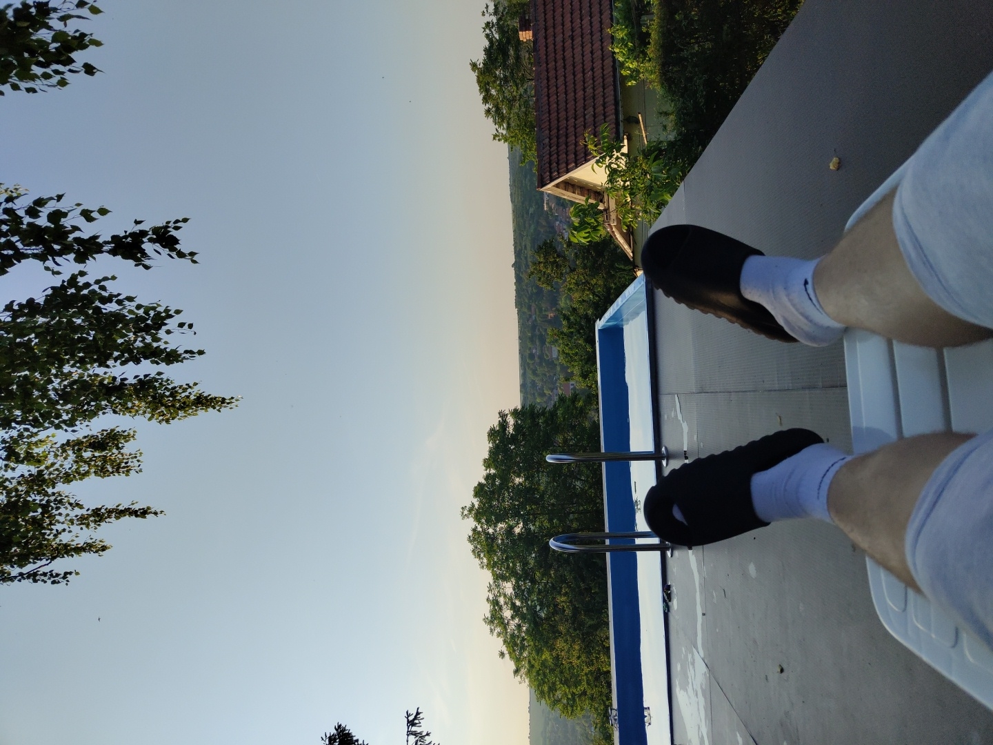 The image captures a first-person perspective from a relaxed, seated position, overlooking an outdoor scene with a pool. In the immediate foreground, the lower legs of an individual are visible, clad in light grey shorts or sweatpants, white socks featuring a dark emblem near the ankle, and dark, slip-on sandals. The individual is occupying a white plastic lounge chair, a portion of which is visible at the bottom center of the frame.
Directly ahead and slightly to the left, a rectangular, above-ground swimming pool is present, featuring a light blue interior and a lighter blue border. Two chrome-finished handrails are positioned at the edge of the pool, extending into the water. The flat surface surrounding the pool and supporting the lounge chair is dark grey or black, appearing to be a composite deck or roof material, with visible water droplets near the pool's edge.
Beyond the pool, a landscape of dense green trees forms the midground and extends into the background, suggesting a heavily vegetated or forested area. To the right of the pool, a section of a residential structure with a reddish-brown tiled roof is partially visible, nestled among the trees. The distant background reveals a sprawling panorama of a settlement or village situated on undulating hills, composed of multiple buildings under a clear, pale blue sky. Overhanging green foliage from a tree drapes into the top portion of the frame. The scene is illuminated by natural light, consistent with daytime. The image provides no specific visual markers to confirm the location as Pisanets, Bulgaria.