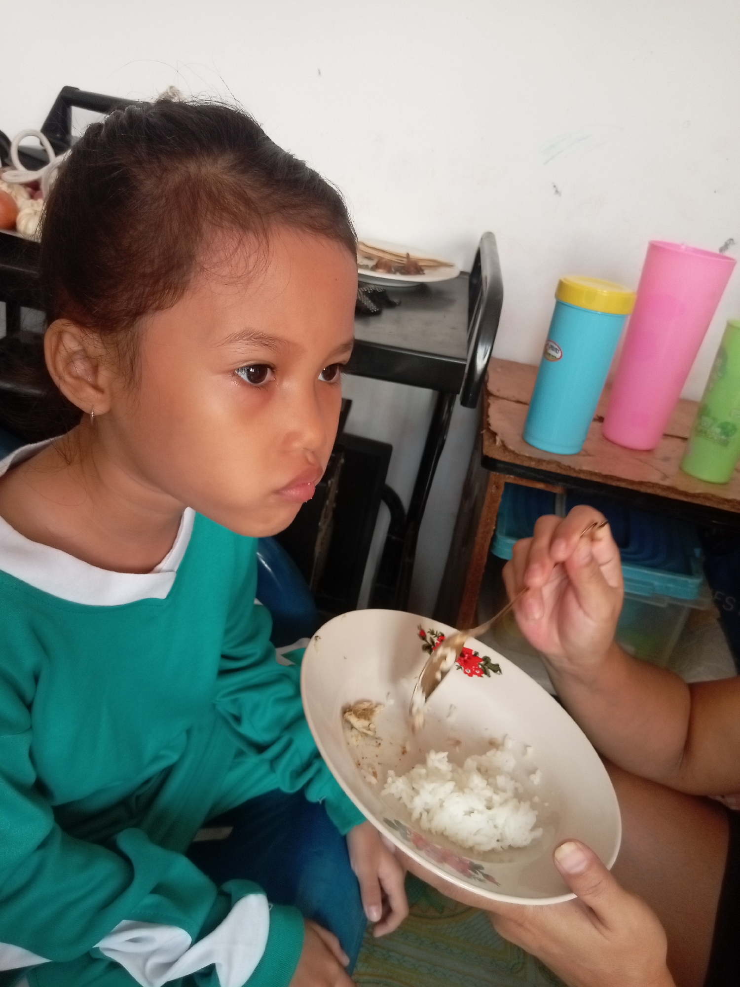 A young Indonesian girl with dark hair tied back is sitting and looking to her right with a somewhat pensive expression. She is wearing a teal long-sleeved shirt with white trim. In front of her, a bowl of white rice and some food is being offered by a hand holding a fork. The bowl has a floral pattern on the rim. The setting appears to be indoors, possibly a home. In the background, there are colorful plastic cups on a shelf, as well as some objects on a dark shelf that could be food items like onions and garlic. There is no visible text in the image. The lighting suggests it is daytime.