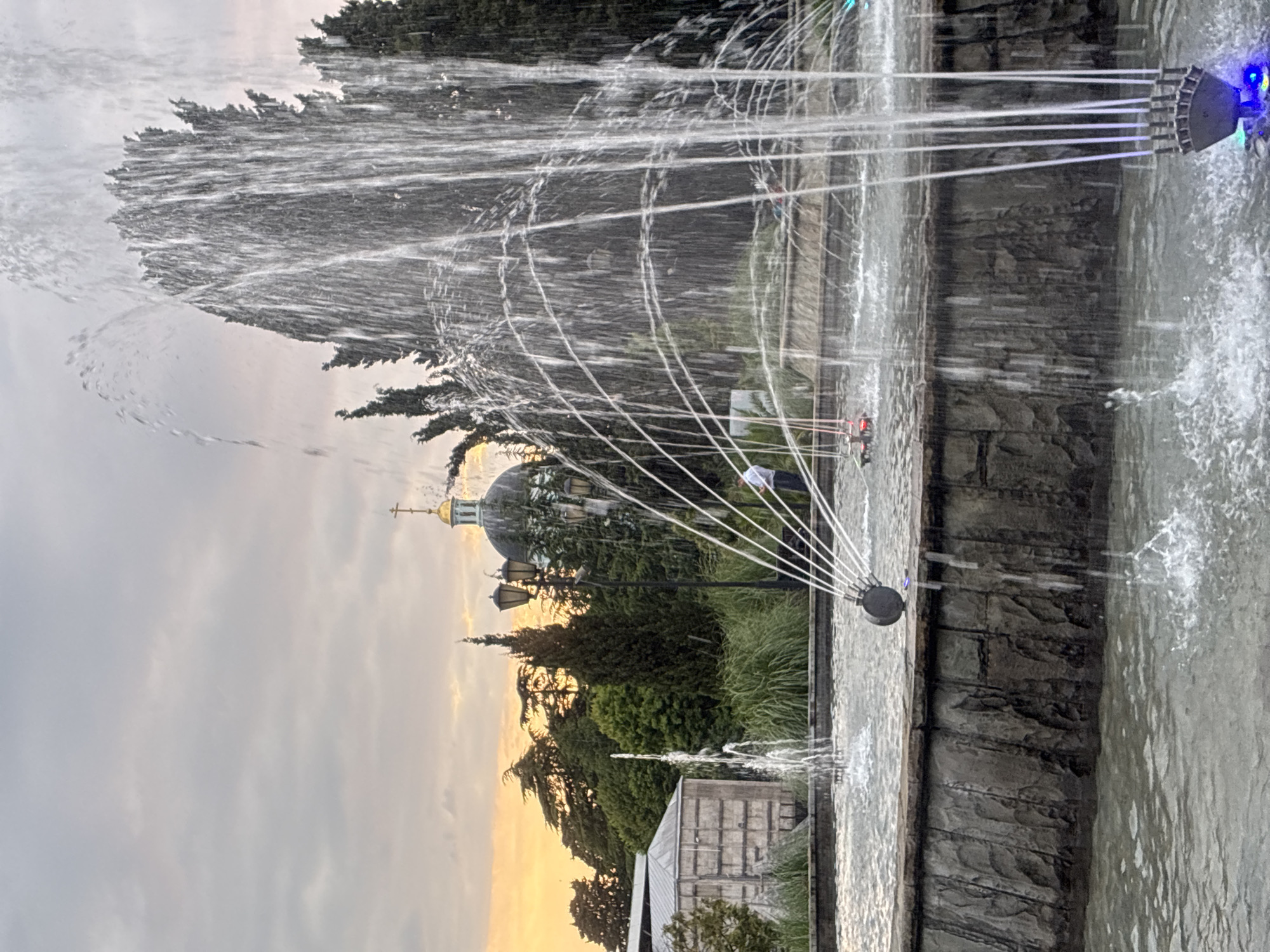 The image depicts a fountain in Sochi, Russia. In the foreground, water sprays from the fountain, creating a dynamic visual effect. The fountain has a stone basin, and some of the water jets are illuminated by blue lights. In the background, a man in a light-colored shirt and dark pants stands near the fountain, observing the scene. To the left, a building with a grey roof is visible, surrounded by lush green trees and vegetation. Further in the background, a domed building with a golden cross on top, likely a church or cathedral, is set against a cloudy sky tinged with the warm hues of sunset. The overall atmosphere suggests a tranquil evening setting in a public park or square.