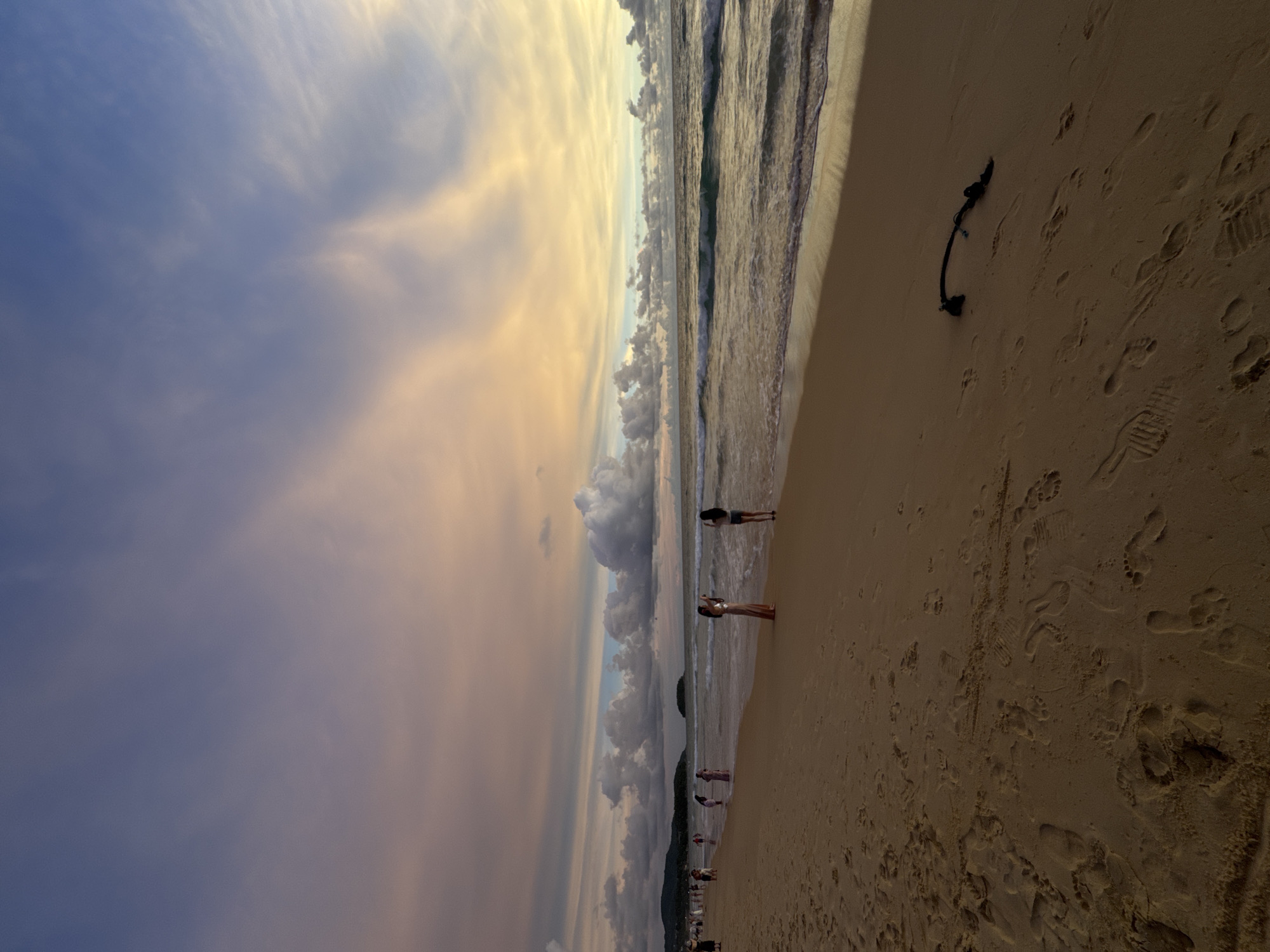 This media file captures a beach scene at Karon, Thailand, likely during sunset or sunrise due to the warm, golden hues in the sky. The image is oriented vertically, with the sky occupying the left two-thirds and the beach and ocean on the right.
The sky is filled with dramatic clouds, ranging from wispy, feathery formations to larger, more defined cumulus clouds. The sunlight filters through, creating a soft, ethereal glow that transitions from a deep blue in the upper left to shades of yellow, orange, and pink towards the horizon. The ocean is calm, with gentle waves lapping the shore.
On the sandy beach, several people are visible. Two women are standing near the water's edge, appearing to be enjoying the view. Further down the beach, more individuals are scattered, some walking and others seemingly relaxing. The sand is marked with numerous footprints, indicating recent activity. A dark, coiled object, possibly a piece of rope or debris, lies on the sand in the foreground. The overall atmosphere appears peaceful and serene.