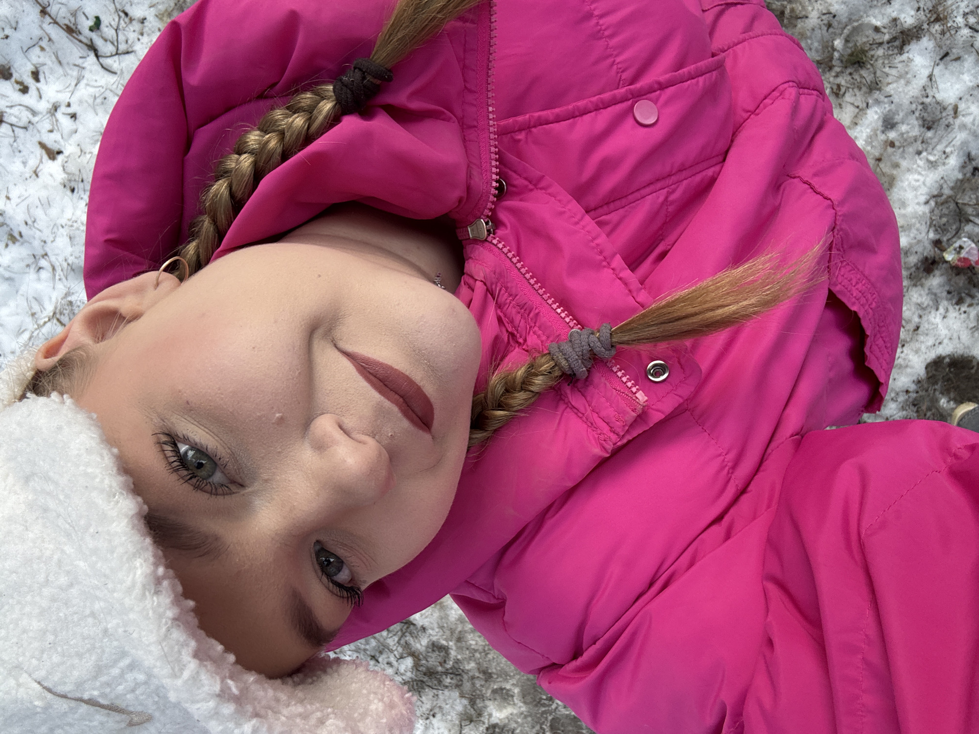 A high-angle photograph depicts a female subject outdoors in the city of Chaikovsky, Russia, against a snowy ground cover. The subject, positioned centrally, is wearing a white fuzzy winter hat with a partially visible dark logo on the front, and a fuchsia-colored puffer jacket. Her light brown hair is styled into two braids, secured with grey hair ties, resting on her shoulders and jacket. She has light-colored eyes (appearing green-grey), well-defined eyelashes, and her lips are covered in a dark matte lipstick. A small gold hoop earring is visible in her left ear. The puffer jacket is zipped, featuring a silver zipper pull and a circular pink snap button on the right chest. The background consists of a patchy layer of snow mixed with dark brown ground debris, including twigs and foliage. The subject is looking directly at the camera with a neutral expression.