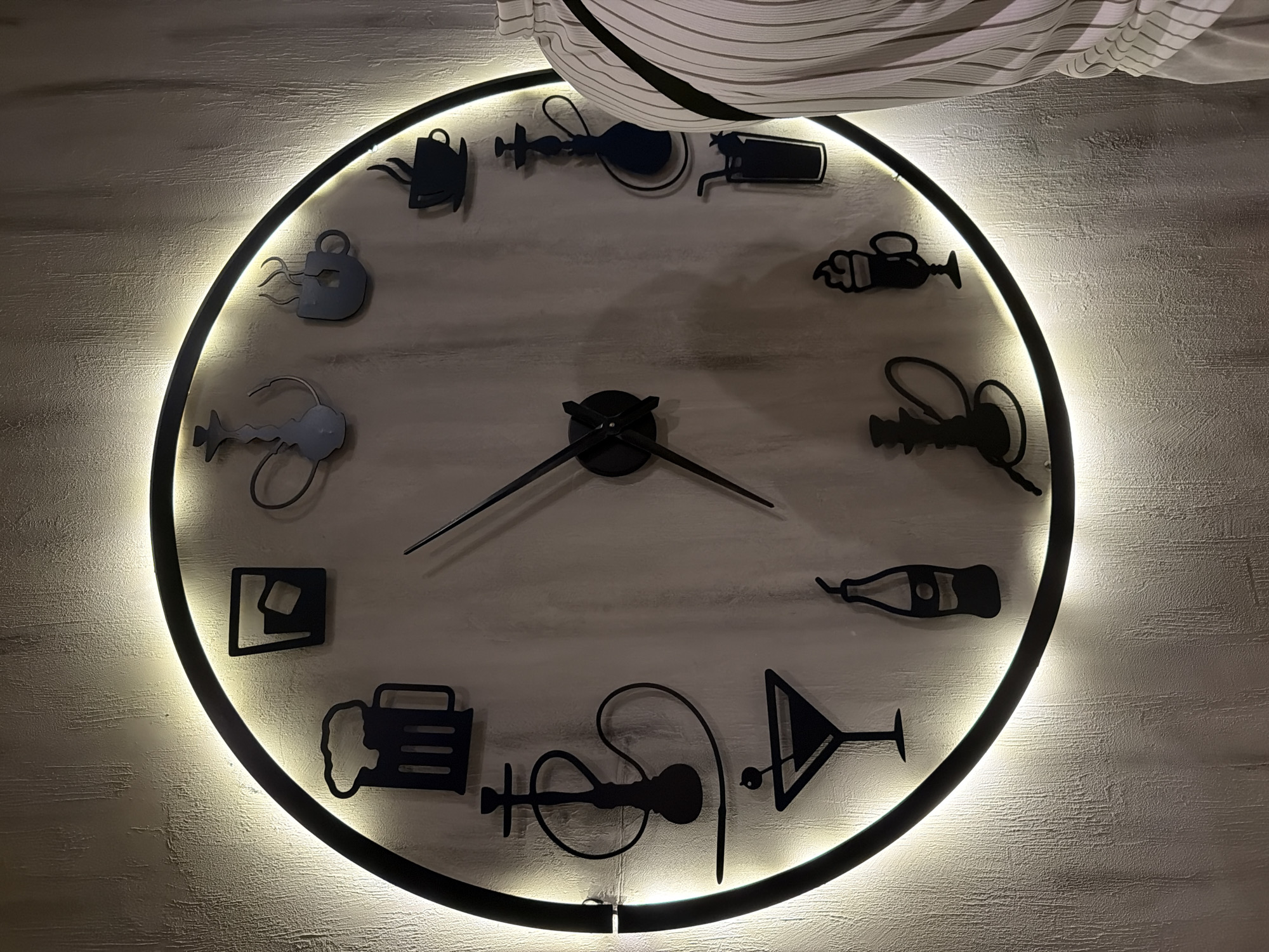 A large, circular wall clock with a black frame and warm white LED backlighting is prominently displayed against a textured, light-colored wall. Instead of traditional numbers, the clock features twelve distinct black silhouettes of various drinking vessels and related items, arranged clockwise around its perimeter.
Starting from the approximate 11 o'clock position and moving clockwise, the markers include: a steaming kettle/teapot, a hookah, a tall glass/mug, a stemmed cocktail glass, a wine/champagne bottle, a martini glass, a different style of hookah, a beer mug, a tumbler glass with ice cubes, a small stemmed glass, another steaming kettle/teapot, and a steaming mug. Black clock hands are centrally placed, indicating approximately 2:40 (the hour hand points toward the 2 o'clock marker, and the minute hand toward the 8 o'clock marker).
A section of light-colored, striped fabric, possibly a curtain or drapery, is visible at the top of the image, partially obscuring the top edge of the clock. The bright backlighting creates a halo effect around the clock, illuminating the textured wall. The thematic design of the clock suggests a setting such as a bar, cafe, or lounge in Sofia, Bulgaria. No text is visible in the image.