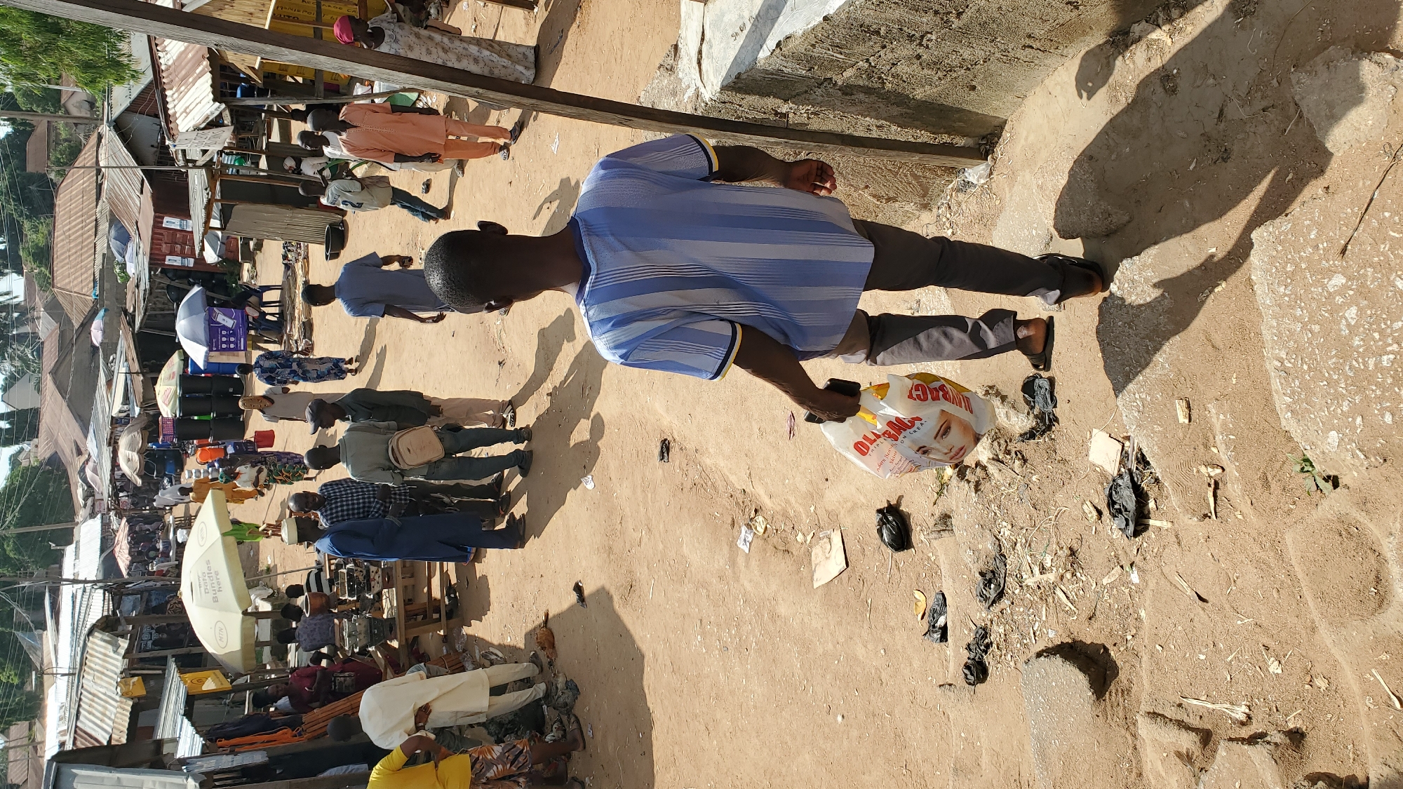 The image depicts a bustling outdoor market scene in Jalingo, Nigeria. In the foreground, a young man wearing a blue and white striped t-shirt and grey pants walks down a dirt path with concrete steps, carrying a plastic shopping bag with an image of a woman's face on it. The path is littered with trash, including black plastic bags and debris. Behind him, the market is crowded with people browsing stalls covered by corrugated metal and fabric awnings. Various goods are visible, including clothing, household items, and umbrellas. People are dressed in diverse traditional and Western attire. The ground is sandy and dusty, typical of the environment in Jalingo. The overall impression is one of vibrant economic activity in a somewhat unkempt urban setting.