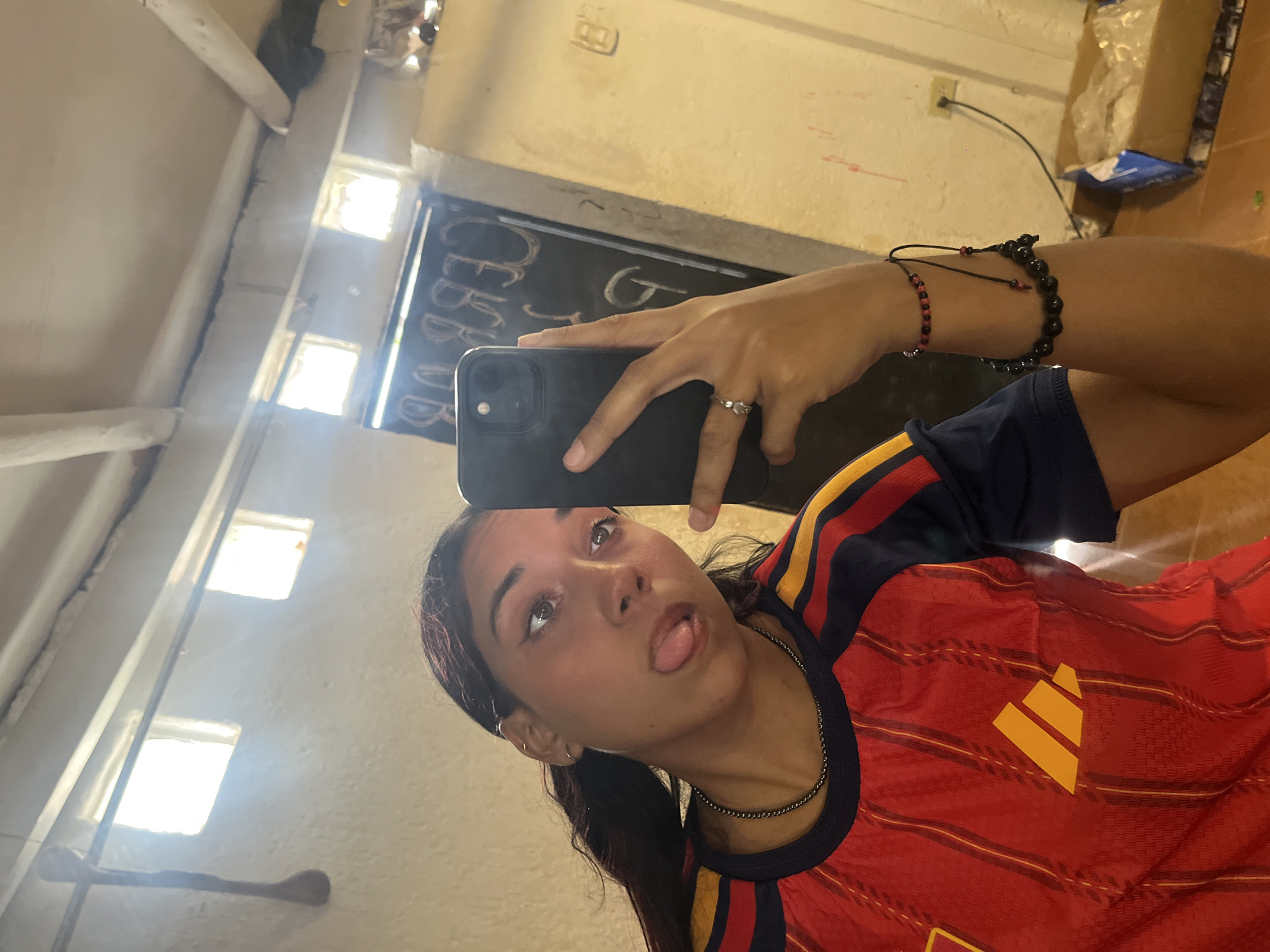 A young woman with dark hair, wearing a red and yellow striped shirt, sticks her tongue out at the camera while taking a selfie in a room. She is wearing a silver ring and several bracelets on her wrist. The room appears to be dimly lit, with a rough concrete wall and ceiling. In the background, a chalkboard with text written on it is visible, along with two small windows and a hanging wooden object. A power outlet and a box are also present. The image was taken in Porlamar, Venezuela.