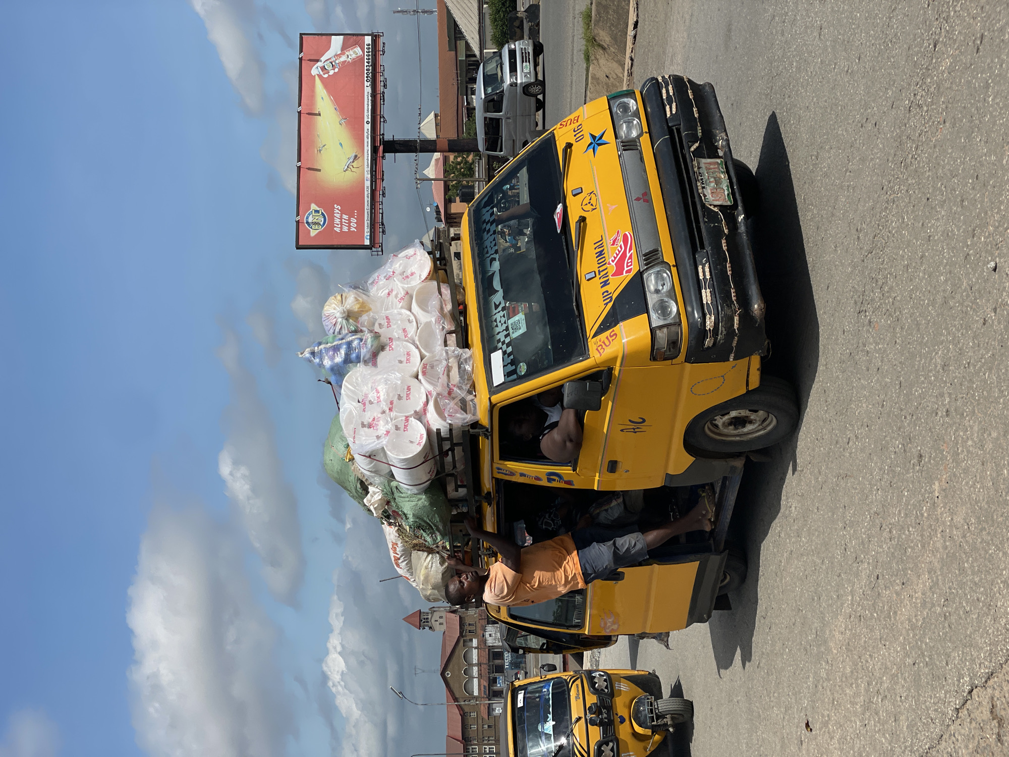 An outdoor scene captured in the city of Unknown, Unknown, under a bright, partly cloudy sky, depicts a bustling street environment. The central focus is a yellow Mitsubishi Fuso L300 van, functioning as a commercial passenger vehicle, heavily loaded with cargo on its roof rack. The cargo includes numerous stacks of white, transparent plastic cups or containers, wrapped in plastic film, along with several large, irregularly shaped sacks, some green and others light-colored.
A man, dressed in an orange t-shirt and dark shorts, stands on the passenger-side step of the yellow van, gripping the edge of the roof rack, actively engaging with or securing the overloaded cargo. He is barefoot or wearing very light footwear. Inside the van, a driver in a white tank top is visible behind the steering wheel, and another individual is partially visible within the vehicle. The van's front features "UP NATIONAL BUS" text, a red shoe graphic, and a blue star sticker, with its black bumper adorned with a string of white LED lights. The license plate "ABS-747XA" is partially readable.
To the immediate left, the front section of a yellow motorized tricycle (Keke Napep type) is visible, also implying public transport. Further down the road, to the right, a silver minivan is seen moving or parked.
In the background, a large billboard with a red face advertises "BNC" insecticide, featuring a graphic of cockroaches being sprayed, and includes the slogan "ALWAYS WITH YOU..." along with social media handles and a phone number (09083466666). Distant buildings, including one with a distinctive red-roofed tower, are visible against the horizon. The asphalt road surface shows signs of wear. Overhead power lines and utility poles are also present, indicating an urban or semi-urban setting.