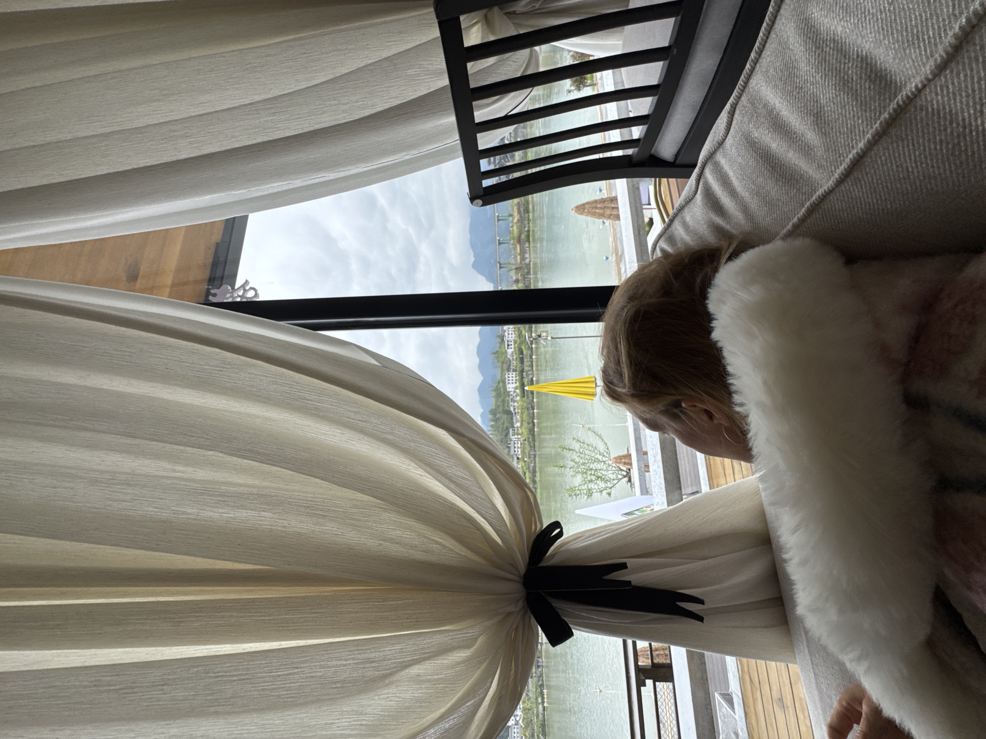 A view from inside a building with a window overlooking an outdoor scene in Zhuoshui, China. A light-skinned person is resting on a light-colored sofa or daybed inside, partially obscured by a white fur-trimmed garment. She has short blonde hair, and her ear is visible. A sheer, light-colored curtain, tied with a black ribbon, drapes across the left side of the frame and around the window. Outside, a body of water is visible, with a bridge spanning across it. Trees and buildings are present along the water's edge. A yellow umbrella is slightly to the right of the bridge, partly in the water and on the bank. Overcast conditions prevail, and mountains are visible in the distance. A dark-colored chair or structure with slats is positioned near the sofa.