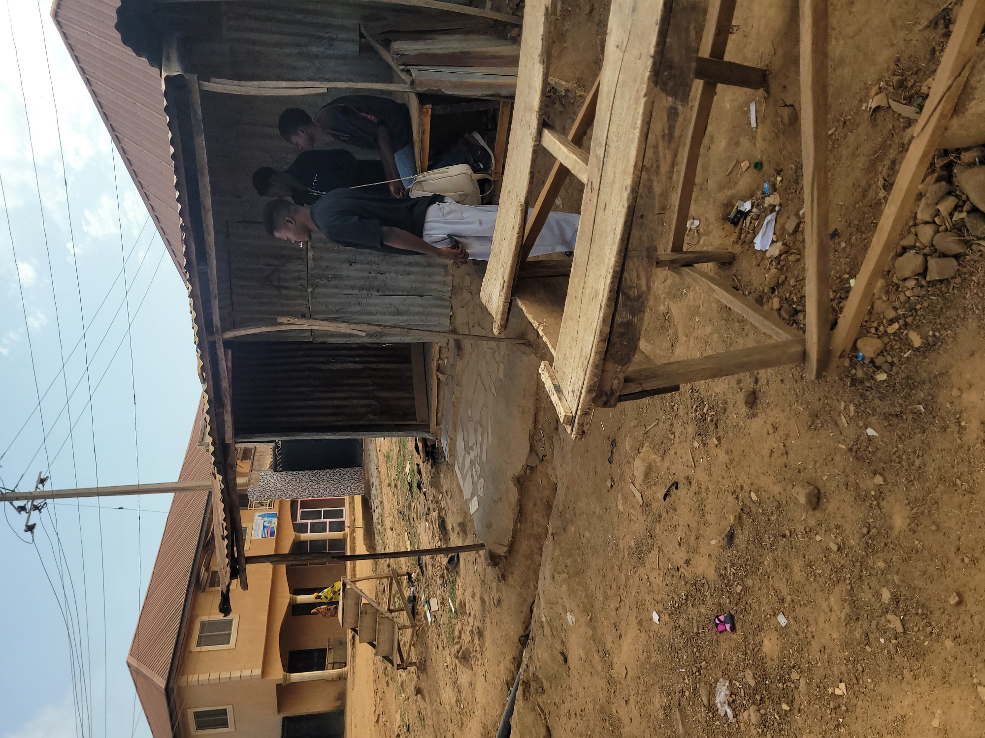 The image captures an outdoor scene in what appears to be a semi-urban or rural setting in Barakin Akawo, Nigeria, under a bright, clear sky. The ground consists of reddish-brown dirt, mixed with pebbles and scattered debris.
In the foreground and to the left, there is a simple, open-fronted structure constructed from corrugated metal sheets for the roof and walls, supported by wooden beams. Inside this shaded area, three young men are gathered. One man, wearing a dark shirt and light-colored pants, is partially visible, appearing to be standing or leaning. Another man in a dark shirt is seen further inside, and a third man's head is visible, looking downwards. A cream-colored handbag is also seen within the structure.
To the right, a rustic wooden table or bench, roughly constructed from planks and beams, occupies part of the foreground. The ground around this structure is littered with various small pieces of trash, including plastic remnants, paper, and a pink and black sandal. A black hose or pipe runs across the dirt ground in the mid-distance.
In the background, a more substantial two-story building with a reddish-brown corrugated metal roof and light brown/tan walls is visible. It features several rectangular windows and some decorative tiling on its upper section. A utility pole with multiple overhead wires stands prominently against the bright sky, extending across the scene.
The scene suggests a casual gathering, with the men likely socializing or resting in the shade during daytime. There is a small, unreadable blue and white sign on the background building.