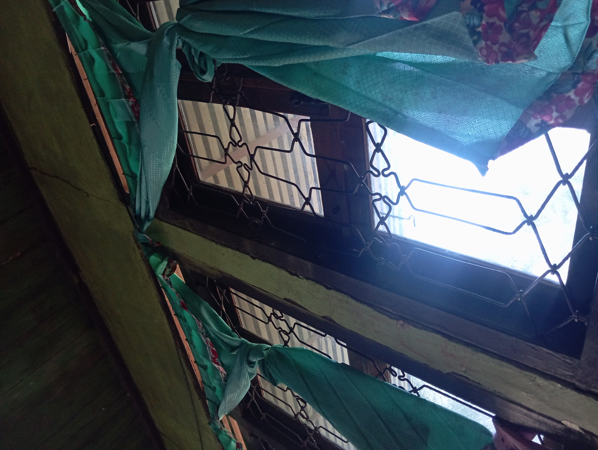 The image shows an interior view of a window in Rejosari, Indonesia. The window features decorative metal bars and is partially obscured by green fabric that is tied in knots. The fabric appears to be a curtain or draped cloth. Sunlight streams through the window, creating a bright area and casting shadows. The overall scene suggests a simple, possibly rustic, setting. The wooden window frame and the weathered appearance of the surrounding wall and ceiling indicate an older structure.