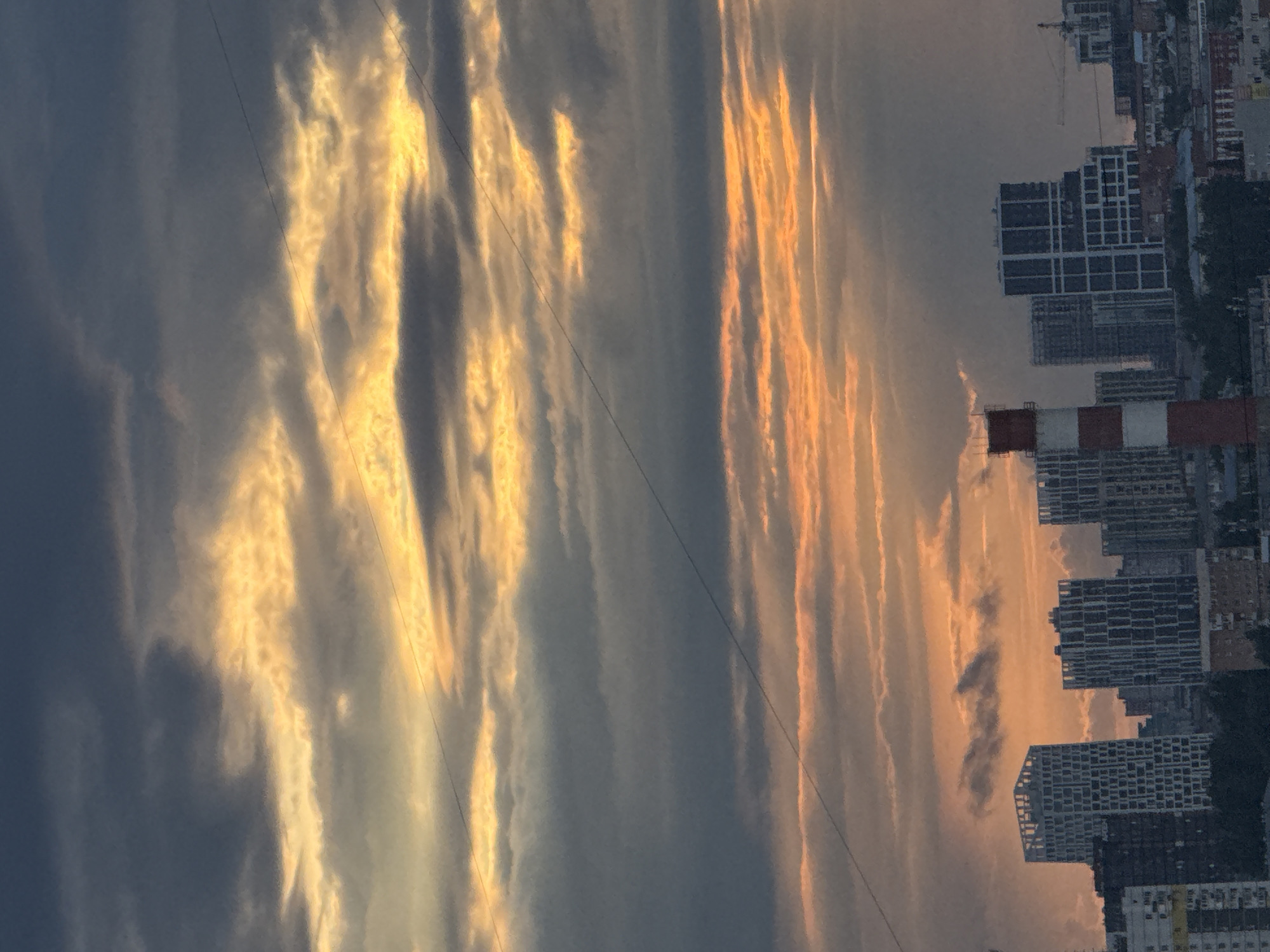 The image captures an urban landscape under a dramatic sky, likely during either sunrise or sunset. The sky dominates the upper two-thirds of the frame, filled with elongated, streaky cloud formations. These clouds are brilliantly illuminated with vibrant golden, orange, and yellow hues, contrasting sharply with the darker, cooler blue-grey tones of the sky and less illuminated clouds behind them. The warm light creates a striking visual effect, highlighting the textures and edges of the clouds.
A city skyline occupies the lower portion of the image. Numerous multi-story buildings, predominantly appearing as residential or commercial structures, are visible. Many of these buildings exhibit a grid-like pattern of windows or architectural elements. In the mid-ground, a prominent industrial chimney or smokestack, painted with alternating red and white stripes, stands out against the sky and surrounding structures. Some buildings appear to be under construction, with exposed concrete frames. Thin, dark power lines are faintly visible stretching across the sky, traversing the cloud formations. There are no people visible in the image. The overall scene conveys a tranquil yet striking atmospheric moment over the city of Izhevsk, Russia, characterized by the dynamic interplay of light and clouds. No text is visible.