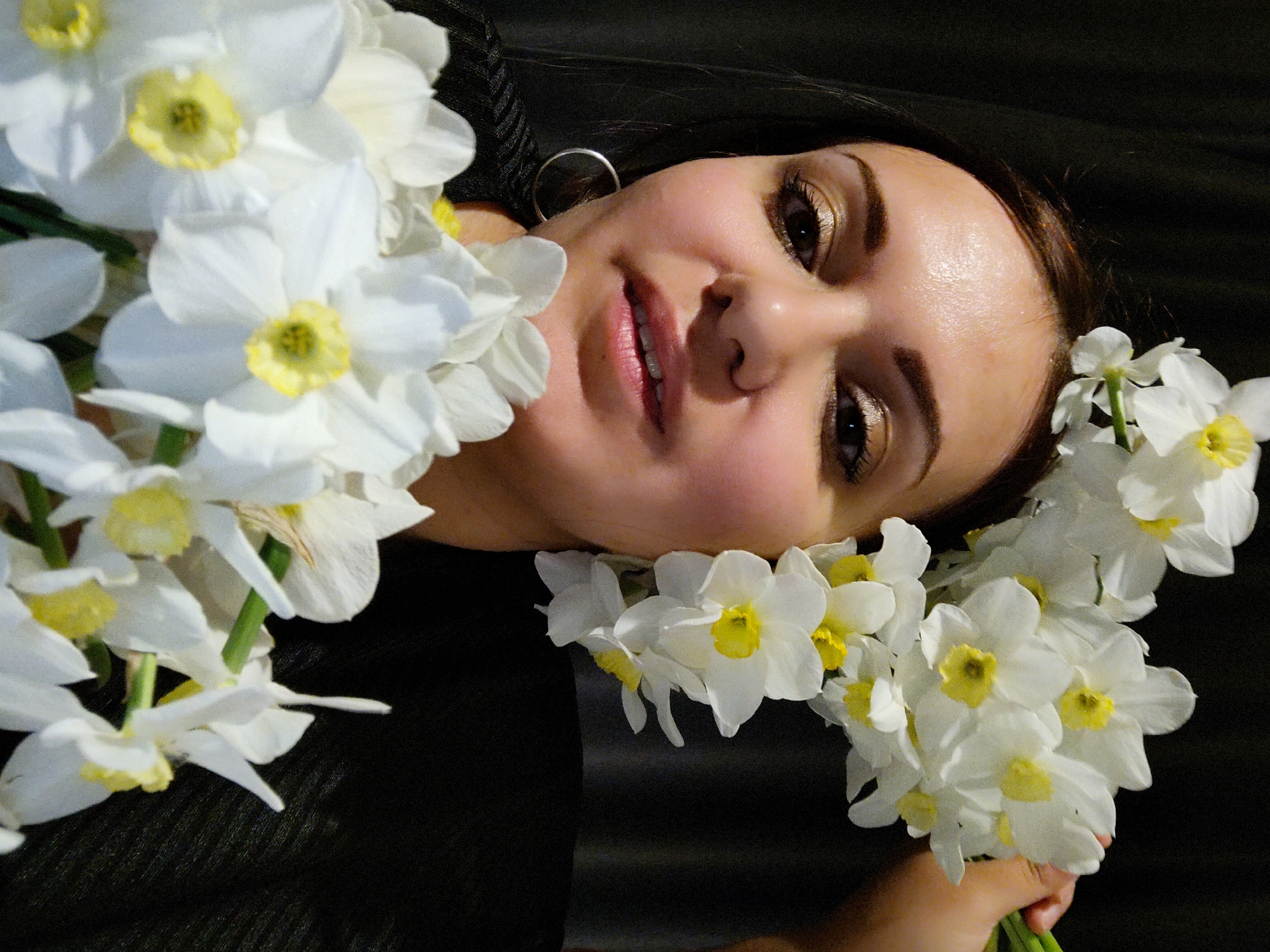 A woman lies on a dark surface with a bouquet of white daffodils. Her face is turned towards the camera. She has fair skin, brown hair, and wears gold-colored eyeshadow. She has an earring in her left ear. It is not possible to confirm the location from the given image.