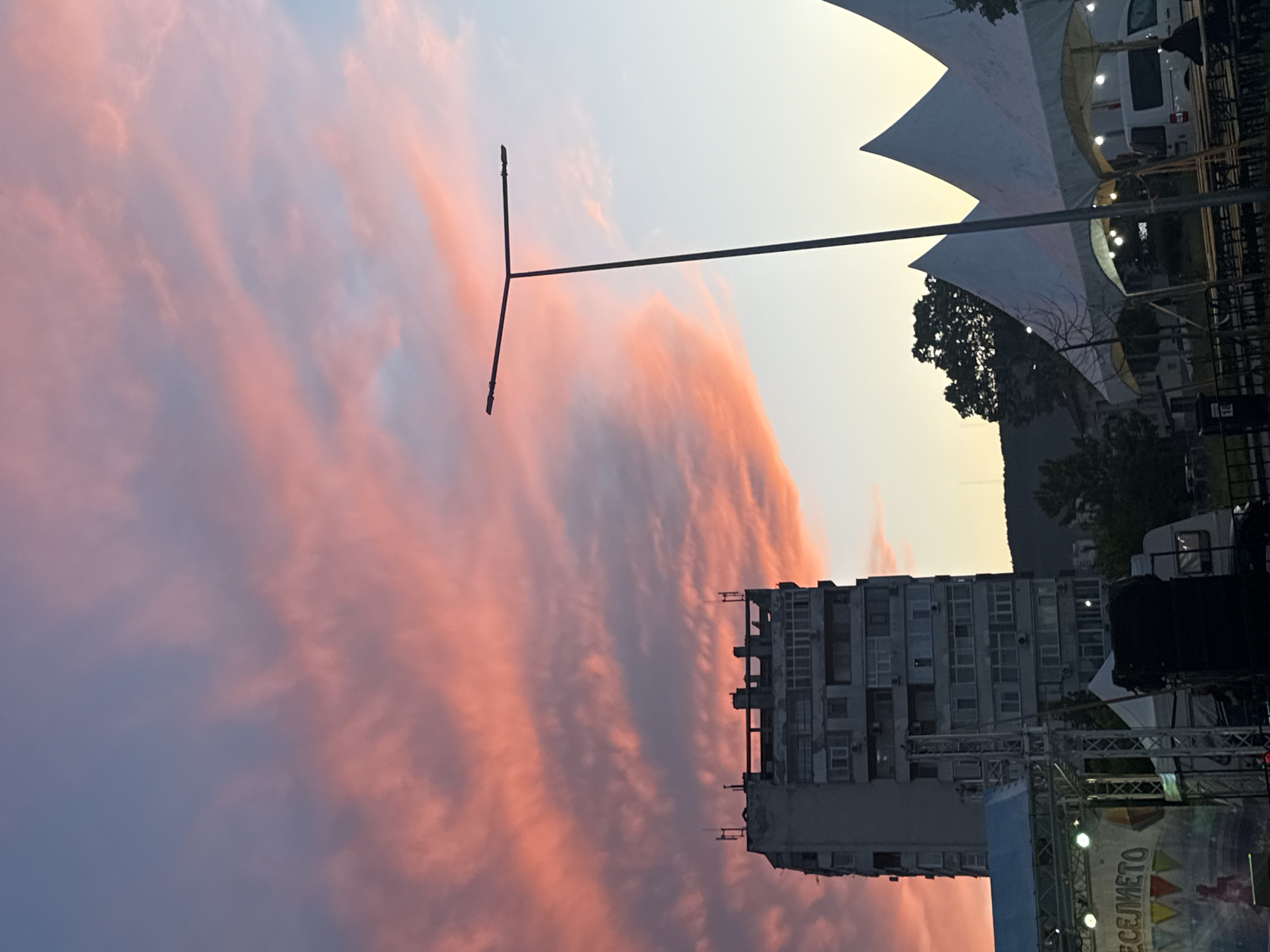 The image captures an outdoor scene in Shumen, Bulgaria, at sunset. The dominant feature is the sky, filled with dramatic, elongated clouds painted in vibrant hues of pink, orange, and purple against a lighter blue and yellow gradient.
In the mid-ground, a tall, dark T-shaped light pole stands out against the colorful sky. To the left (when upright), a multi-story apartment building with numerous windows and balconies is visible, silhouetted against the bright clouds. Parts of the building appear aged or industrial, with some exposed structural elements.
On the right side of the frame, several large, white, tent-like structures with peaked tops are present, suggesting an event or temporary setup. Lights can be seen glowing from beneath these structures. A white van or camper is parked near them, and glimpses of trees and a distant dark hillside add to the natural backdrop.
At the bottom right, elements of what appears to be a stage or event infrastructure are visible, including metal trusses, lights, and a blue section. A banner displays the Cyrillic word "ВЕСЕЛИЕТО," which translates to "The Fun" or "The Merriment," strongly indicating an ongoing or upcoming festival, concert, or public gathering. The overall ambiance is one of evening twilight, setting a mood for an outdoor event. No individuals are clearly discernible in the image.