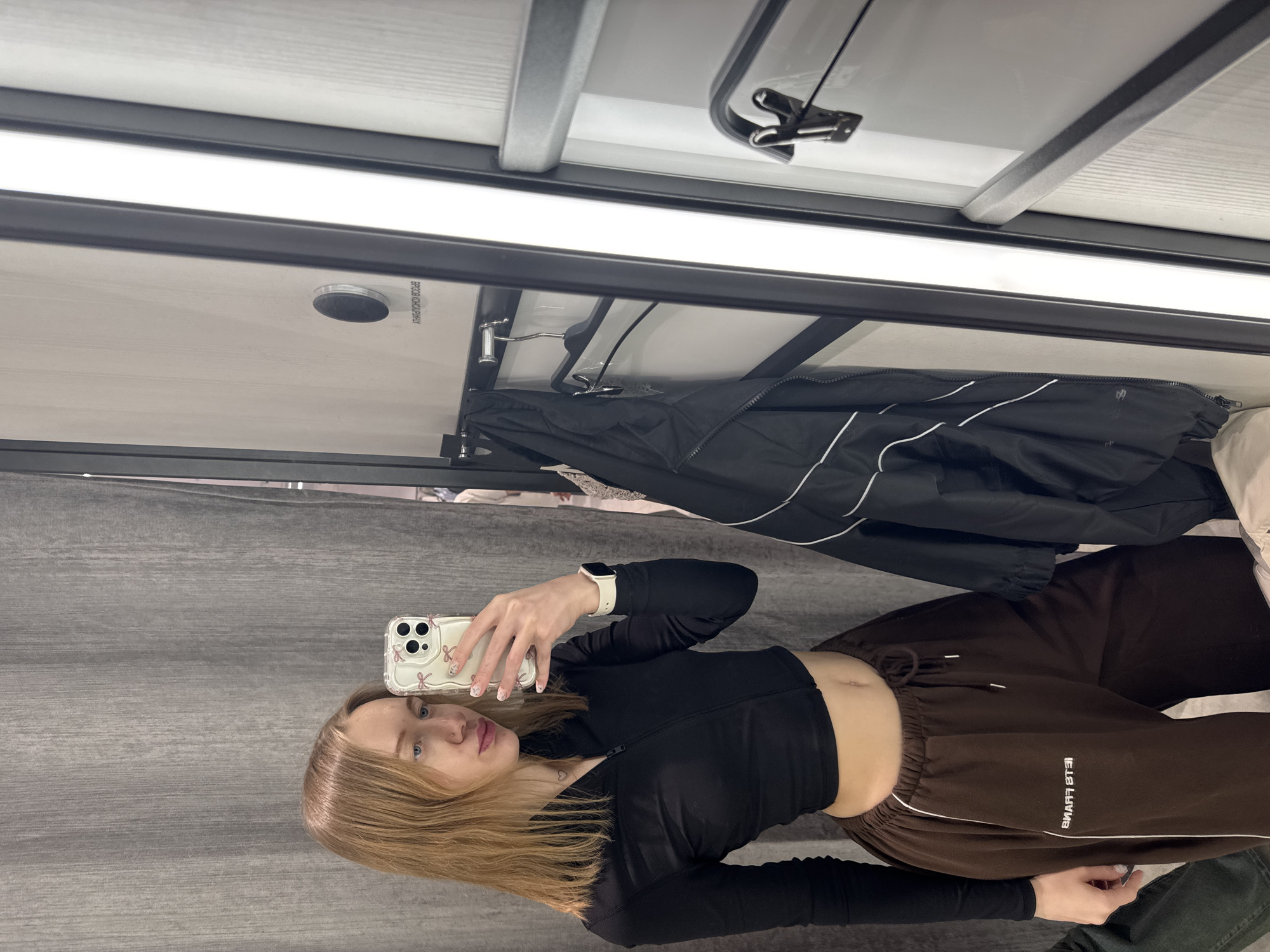 A young woman is taking a selfie in a dressing room. She is wearing a black long-sleeved shirt and brown sweatpants with a white stripe. She is holding a smartphone with a decorative case. A black jacket hangs on a rack behind her. The dressing room has a gray curtain and white walls with a fluorescent light fixture. The city is not mentioned or apparent in the image, nor is there any indication the location is Rostov-on-Don, Russia.
