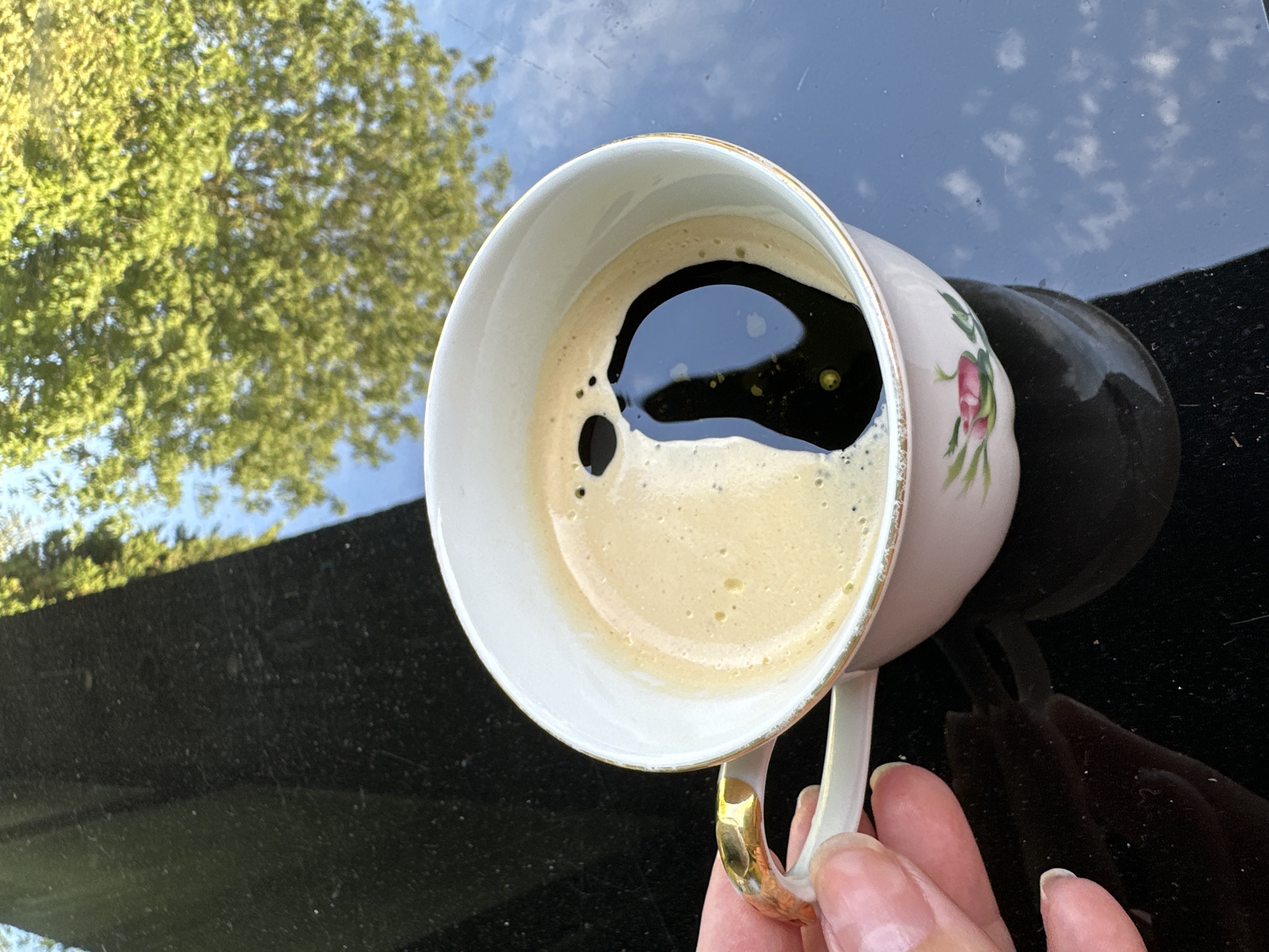 An overhead shot shows a white teacup with a gilded handle filled with coffee. The coffee's foam has created a dark, yin-yang-like shape in the center of the cup. The teacup sits on a reflective black surface, which shows a reflection of trees and sky. The scene is set in Krefeld, Germany, on a sunny day.