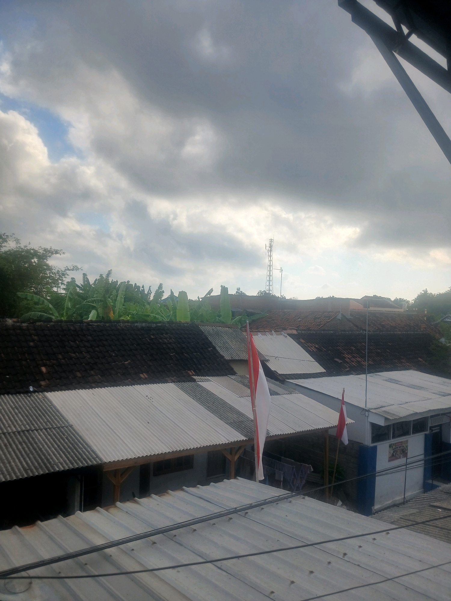 The image captures a view of rooftops and trees under a cloudy sky, possibly in the city of Banyuwangi, Indonesia. In the foreground, corrugated metal roofs are visible. Behind these, a variety of buildings with tiled and corrugated metal roofs are present. To the left, a dense cluster of banana trees forms a prominent green backdrop. Two Indonesian flags, red and white, are displayed on poles. In the distance, a cell tower rises above the tree line. The sky is overcast with a mix of dark and lighter clouds, with a sliver of blue sky visible in the upper left quadrant. The overall impression is of a residential or semi-urban area with significant greenery.