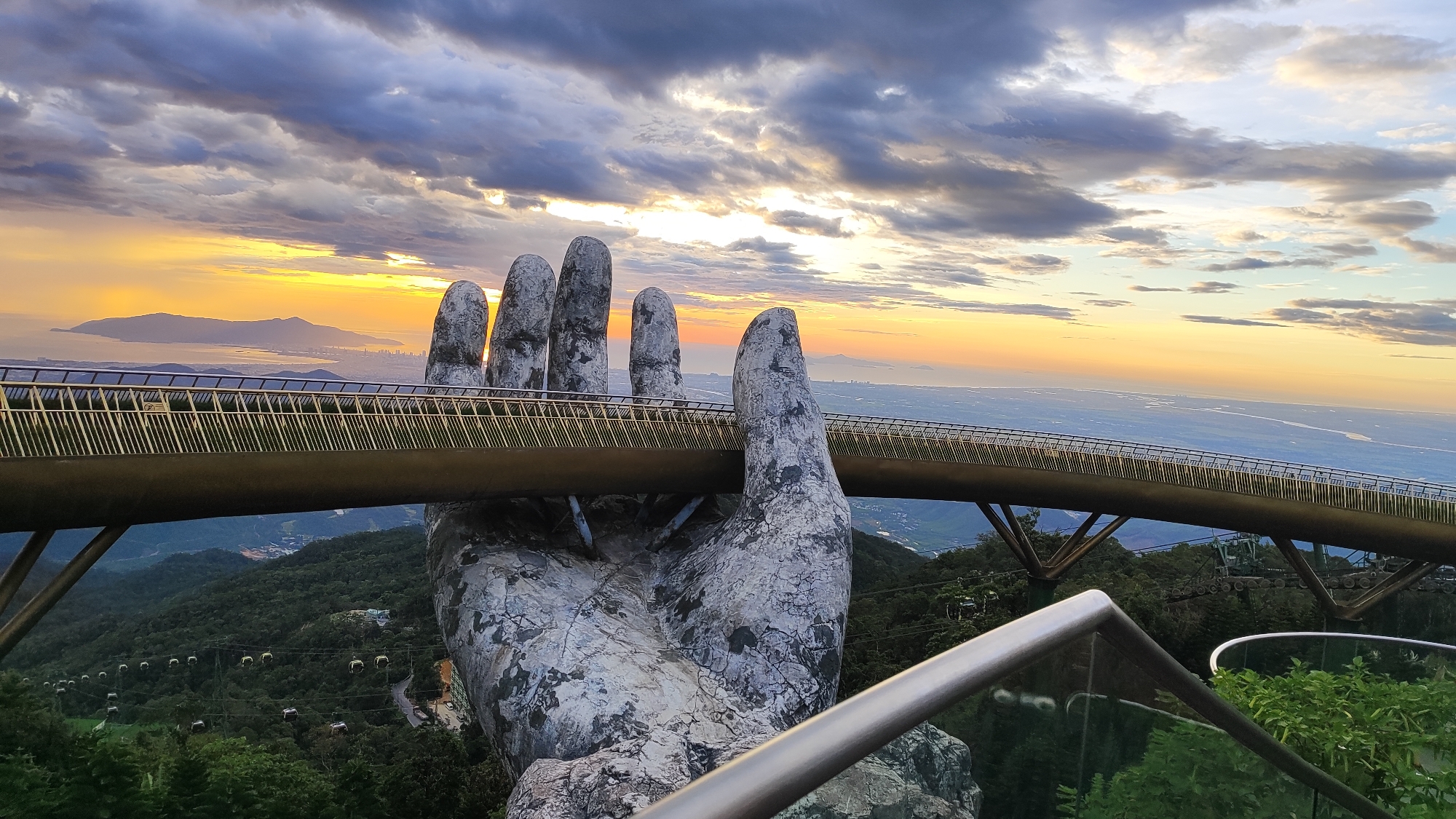 The image shows the Golden Bridge, a pedestrian bridge in the Bà Nà Hills resort in Đà Nẵng, Vietnam. The bridge is held up by two giant stone hands. The scene is captured at sunset, with the sky painted in hues of orange, yellow, and purple. The bridge spans across a lush green valley, with cable cars visible in the distance. The foreground features a metal railing and greenery, suggesting a viewpoint from a higher elevation. The overall atmosphere is dramatic and picturesque, highlighting the unique architecture of the bridge against the natural landscape.