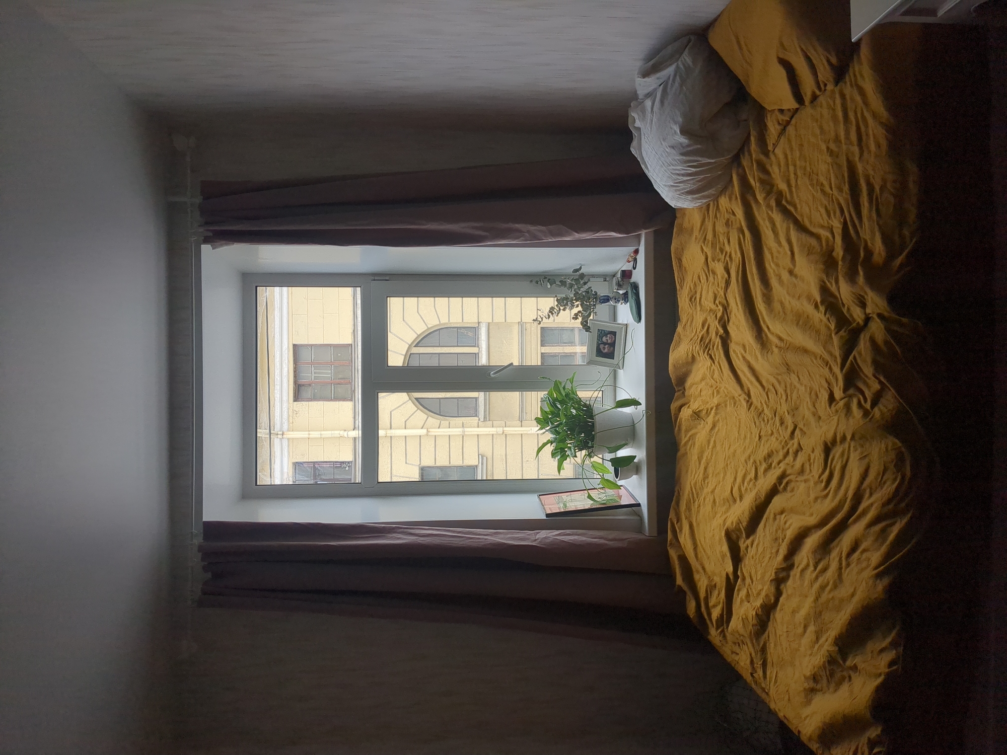 A bedroom contains a window with brown curtains. The window faces a building with classical architecture, likely located in Saint Petersburg, Russia. A bed with a mustard-colored duvet and white pillows is partially visible in the right side of the frame. Plants and a picture frame sit on the windowsill. The room appears dimly lit.