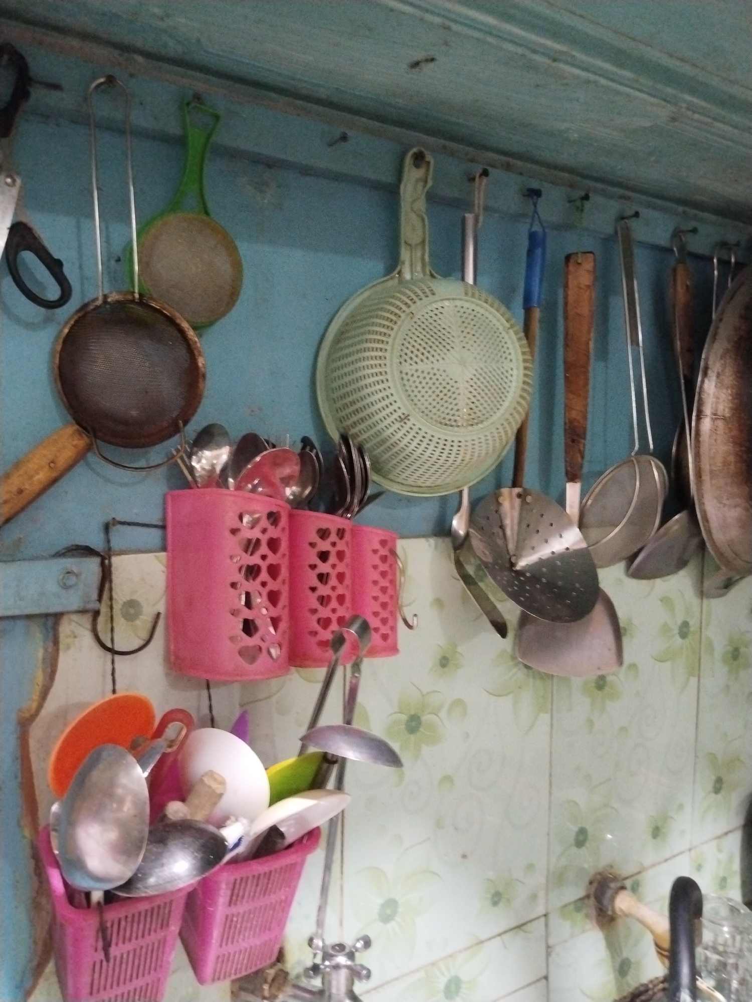 This image depicts a close-up, eye-level view of a collection of kitchen utensils hanging on a light blue wall in what appears to be a residential kitchen in Prigi, Indonesia. The scene is cluttered, with various metal and plastic items suspended from hooks and nails.
Prominently featured are several strainers and ladles, some with perforated metal heads, hanging in a row. To the left, a metal strainer with a wooden handle is visible, as is a green slotted spoon. Below these, two pink plastic holders, shaped like small baskets with heart cutouts, are filled with spoons and other utensils.
Further to the right, a large, light green colander with a wide rim is the most eye-catching item. Beneath it, more metal ladles and strainers are arranged. A large, dark-colored pot or pan hangs on the far right edge of the frame.
The wall behind the utensils is painted a light blue, with some wear and tear visible. Below the utensils, a section of tiled wall with a light green floral pattern is visible, indicating the lower part of the kitchen backsplash. A metal faucet with a wooden handle is partially visible at the bottom right, suggesting a sink area nearby. The overall impression is of a functional, well-used kitchen space.