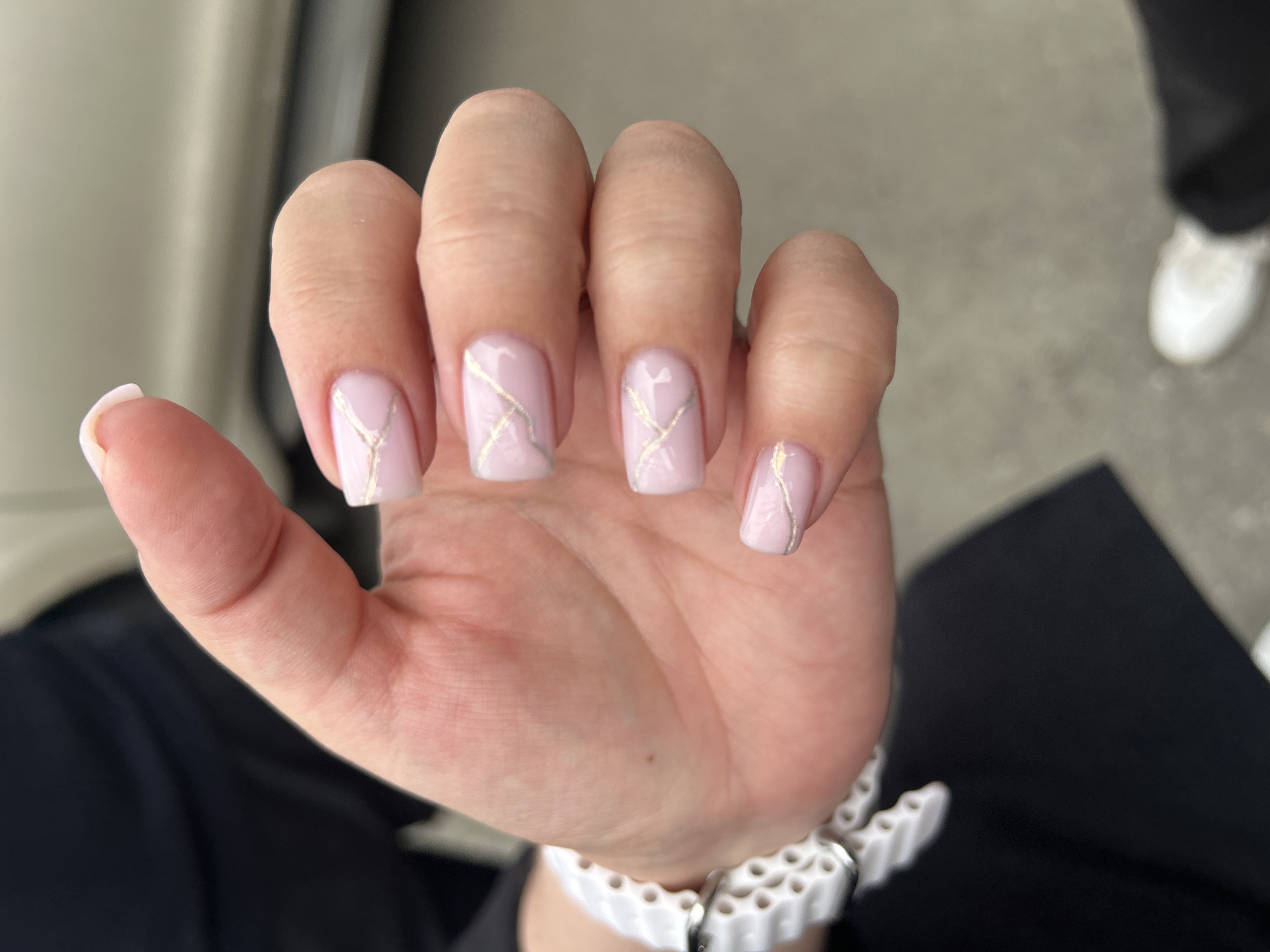 A close-up, eye-level, vertical shot shows a person's left hand, which is adorned with light pink, square-shaped fingernails decorated with thin, irregular gold lines. The hand is positioned with the palm facing downwards and slightly towards the viewer, revealing a white, perforated watch on the wrist. The background is out of focus, showing the blurred shapes of black clothing and what appears to be a light-colored vehicle. The ground beneath is a neutral gray, with a white sneaker visible in the lower right corner of the frame. The lighting suggests an outdoor setting during the daytime. No specific landmarks or elements within the image suggest the city of Perm, Russia.