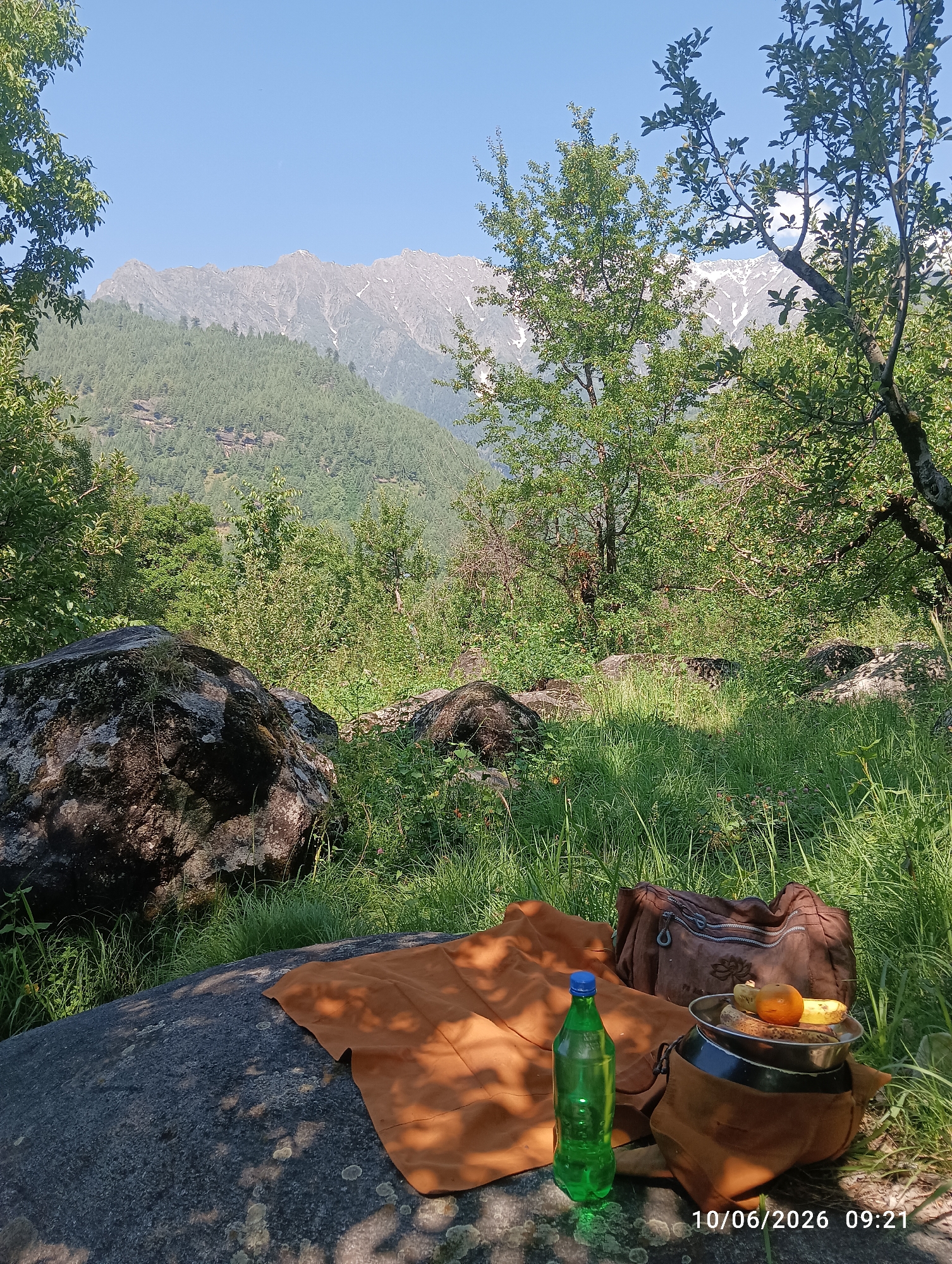 A picnic setup is arranged on a large, dark grey boulder in a grassy, forested area. In the foreground, an orange cloth is spread over the boulder, next to a green plastic water bottle. Behind the bottle, a brown woven bag and a bowl filled with fruit, including bananas and an orange, are placed on top of a dark brown cloth. The background features lush green trees and distant, snow-capped mountains under a clear blue sky. The timestamp "10/06/2026 09:21" is visible in the bottom right corner of the image. The scene is peaceful and appears to be a scenic spot for outdoor relaxation. There is no information provided to indicate this location is in Barheta, India.