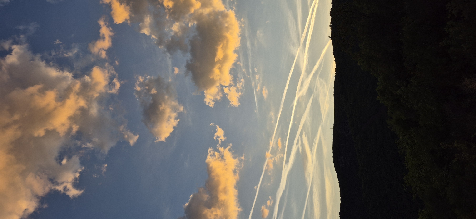 The image shows a daytime sky over Boreti, Montenegro. Orange-tinged clouds are visible in the blue sky, alongside multiple white contrails. A dark, tree-covered hillside is visible on the right edge of the image.