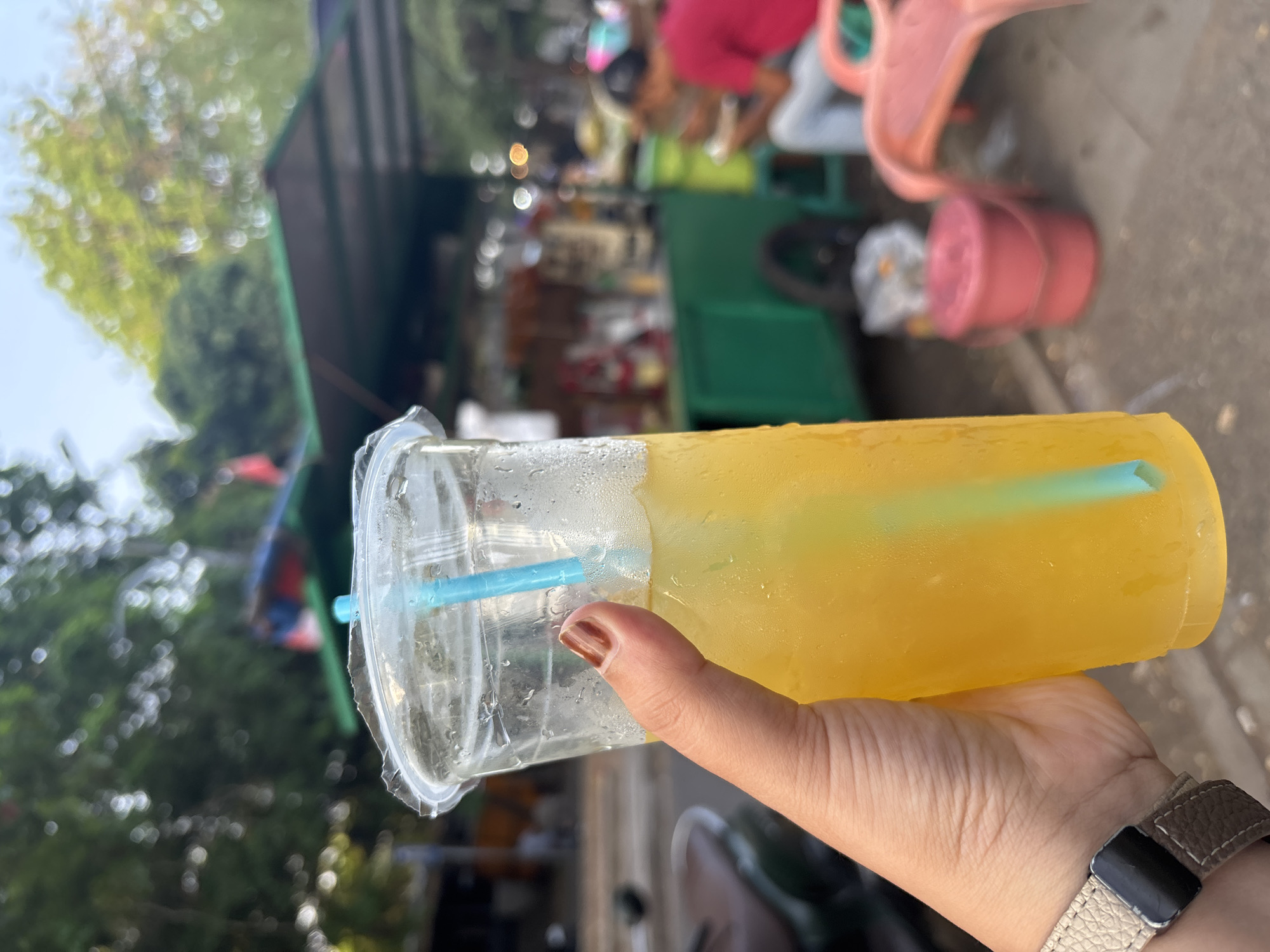 A hand is holding a clear plastic cup filled with a bright yellow beverage and a blue straw. The cup is sweating, indicating a cold drink. In the background, there is a green food cart with various items displayed and a person wearing a red shirt and blue jeans seated on a pink plastic chair. The scene appears to be outdoors, possibly on a street in Cirebon, Indonesia, with trees and foliage visible. The overall atmosphere suggests a casual, outdoor setting.