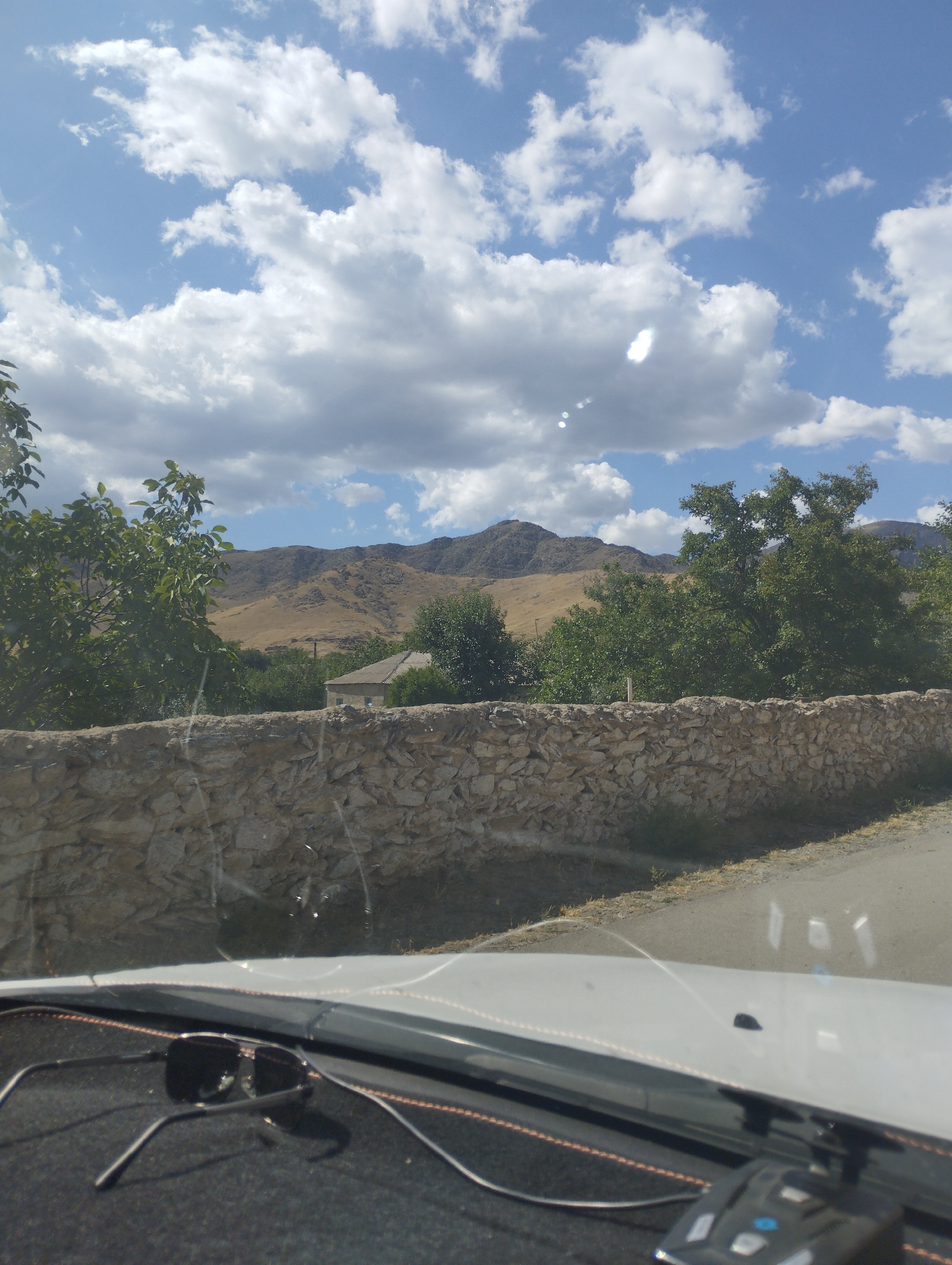 The image shows a view from inside a vehicle, looking out towards a rural landscape in O'xum QFY, Uzbekistan. A stone wall runs horizontally across the mid-ground, with trees and vegetation behind it. A house with a sloped roof is visible nestled among the trees. In the background, rolling hills and mountains are seen under a blue sky with scattered white clouds. The foreground shows a portion of the vehicle's dashboard, with a pair of sunglasses resting on it. The overall scene depicts a sunny day in a rural setting.