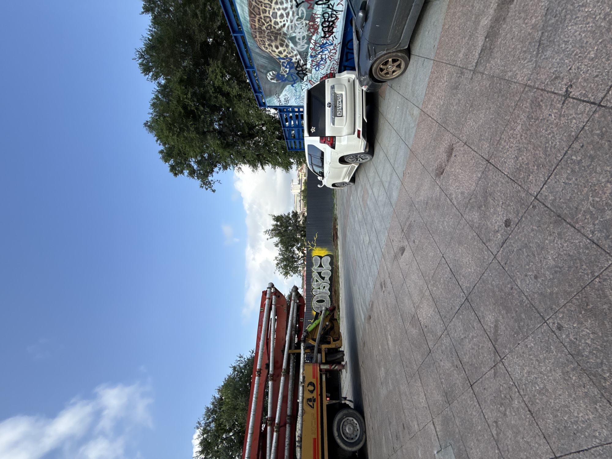 A white Toyota Land Cruiser Prado with license plate A269YM125 is parked on a paved area. To its right, a dark-colored car is partially visible. Behind the Toyota, a large blue structure displays graffiti and a mural of a leopard. A red concrete pump truck with the number "40" is parked to the left of the Toyota. The ground is paved with large square tiles. In the background, trees and buildings are visible under a blue sky with scattered clouds. The location is identified as the area near the Funicular in Vladivostok, Russia, based on visual cues and context from similar images.