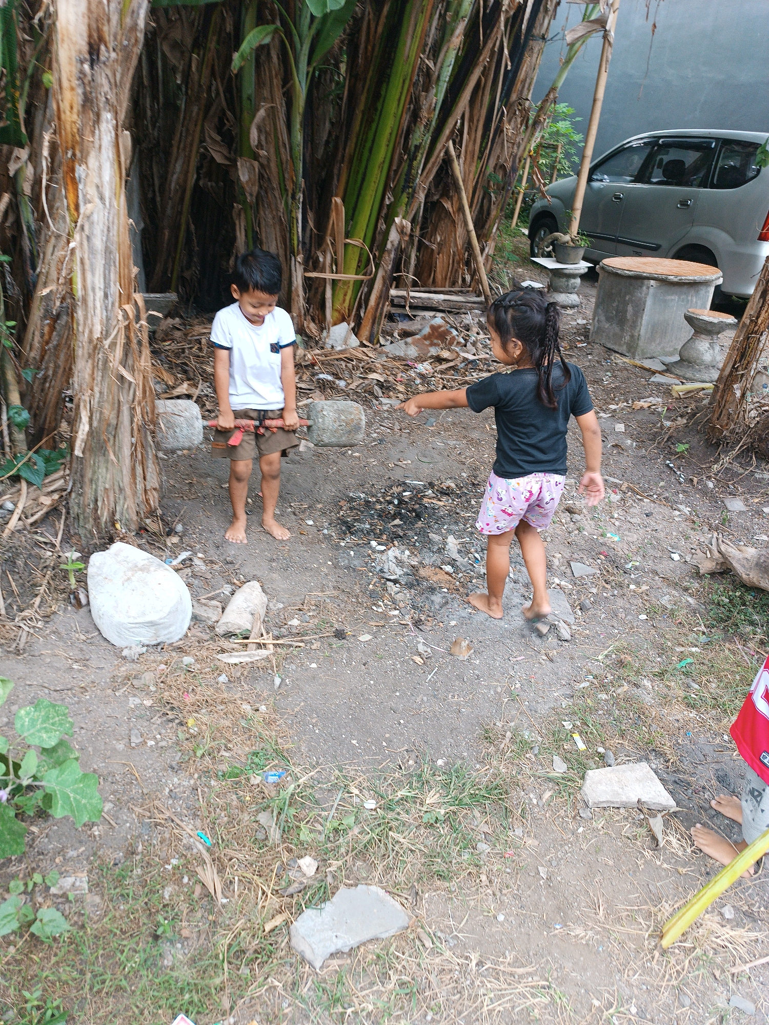 In Tabanan, Indonesia, two children are engaged in outdoor play amidst a rustic environment. A young boy, wearing a white t-shirt and brown shorts, stands barefoot, holding a makeshift barbell constructed from two large stones and a red bar. Opposite him, a young girl, dressed in a black t-shirt and pink patterned shorts, points towards him with her left hand. The scene is set in a somewhat cluttered yard, with dried leaves, debris, and sparse vegetation covering the ground. In the background, a silver car is parked, and several large banana trees with their characteristic thick trunks dominate the left side of the frame. Two concrete structures, one resembling a pedestal and another a table, are visible near the car. The overall impression is one of a casual, everyday moment in a semi-rural setting.
