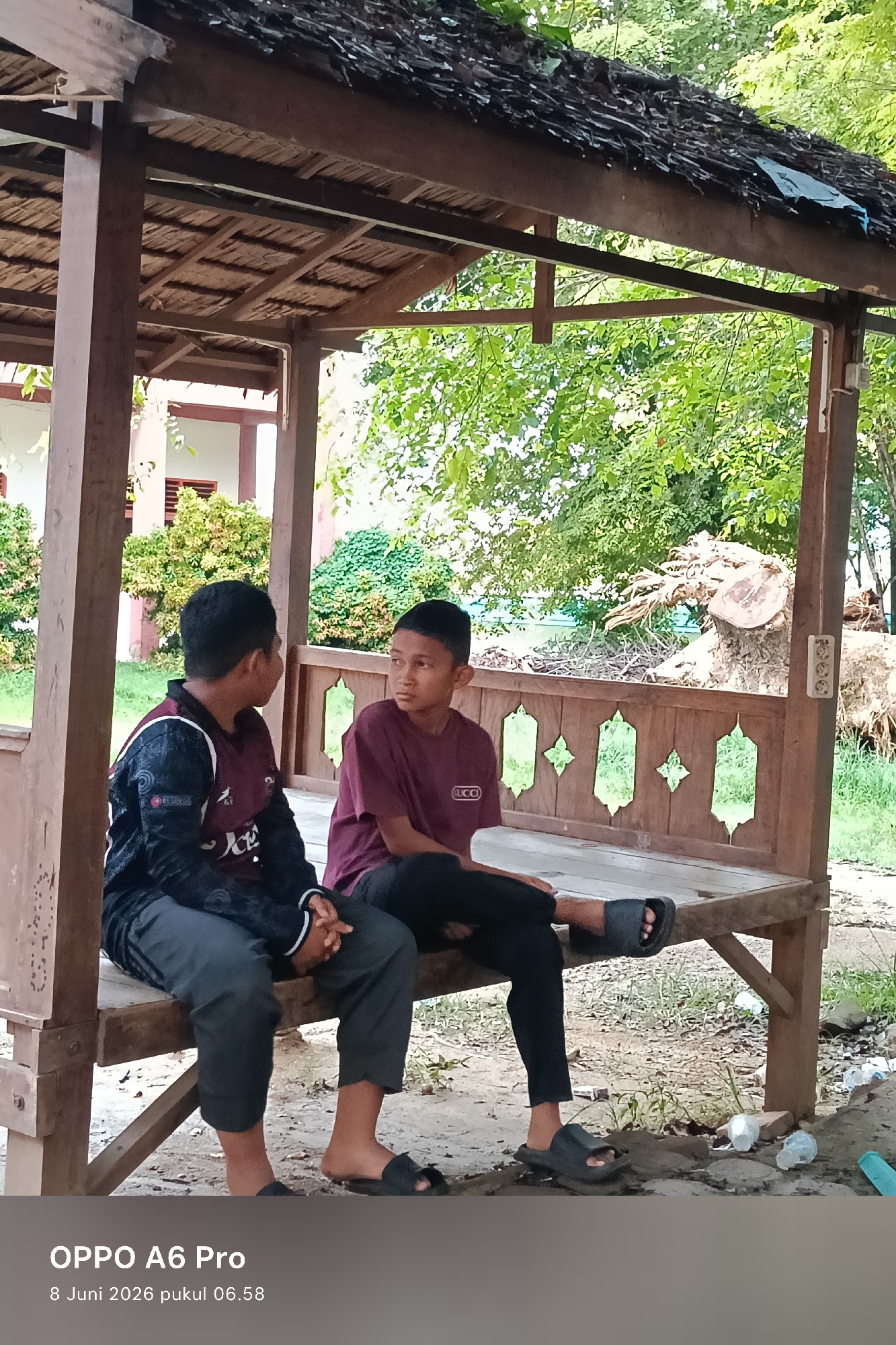 Two young boys are sitting on a wooden bench under a thatched-roof gazebo. The boy on the left is wearing a dark shirt with maroon and white accents and dark gray pants. The boy on the right is wearing a maroon t-shirt and dark pants. They are facing each other and appear to be conversing. The gazebo has ornate wooden railings and a dirt floor. In the background, there are trees, a building, and a large tree stump. The scene is set in an outdoor, possibly rural, area. The timestamp at the bottom indicates the image was taken on June 8, 2026, at 6:58 AM, and is branded with "OPPO A6 Pro." The location is identified as Lhokseumawe, Indonesia.