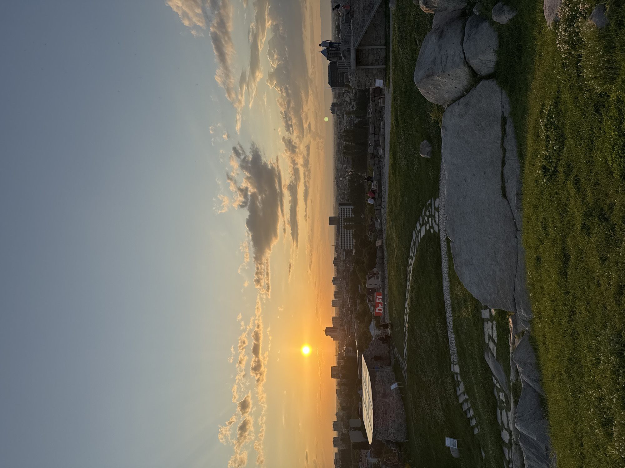 The image captures a panoramic view of a city at sunset, observed from an elevated, landscaped area. The horizon is dominated by a bright, round, yellow-orange sun positioned slightly to the left of the frame's center. The sky transitions from a soft blue at the top to vibrant orange and yellow hues near the sun, with scattered stratocumulus clouds illuminated by the setting light. Several crepuscular rays are visible extending upwards from behind the clouds on the left side of the sky.
Below the horizon, the sprawling cityscape consists of numerous buildings, including multi-story structures and residential areas, extending into the distant haze. The overall impression is one of a dense urban environment.
In the mid-ground, a park-like setting with green grass and large, grey boulders is visible. To the left, a unique stone structure with a curved, possibly metal or corrugated roof stands. Several individuals are gathered around this structure, some sitting. A red sign displaying "Dr. Fl" is positioned near this building. To the right, another ancient-looking stone structure with a modern set of stairs leads up to a flat platform where a person is standing, observing the view. Additional individuals are scattered across the elevated area, some seated on stone walls or benches, enjoying the sunset.
The foreground features lush green grass with scattered small white flowers, and large grey rock formations. A winding path made of irregular flat stones traverses the grassy slope, leading upwards from the lower right. The light from the setting sun casts long shadows, particularly in the foreground, and creates a warm, golden glow across the mid-ground and city.
This scene is identified as being in the city of Plovdiv, Bulgaria, likely from one of its historic hills (such as Nebet Tepe), which offers expansive views of the city and features archaeological elements integrated with modern landscaping.