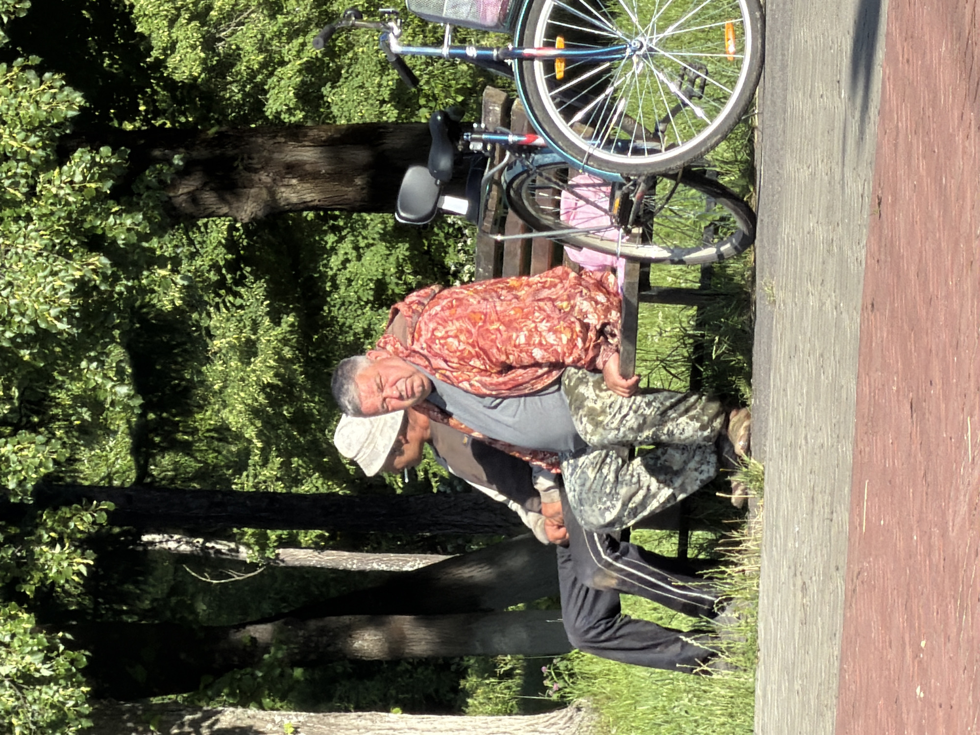 The image shows two men interacting on a sunny day. One man, wearing a colorful patterned shirt, is leaning back and holding onto the other man who is in front of him. The second man is wearing a grey shirt and camouflage pants, and appears to be supporting the first man. Both men are outdoors in what appears to be a park or recreational area, with trees and grass visible in the background. A bicycle is parked nearby, leaning against a bench. The setting suggests a casual, daytime scene. The facial expressions of the men are somewhat obscured, but the overall atmosphere seems relaxed. There is no visible text in the image. The location context of Krasny Oktyabr, Russia, places this scene in a specific geographical area.
