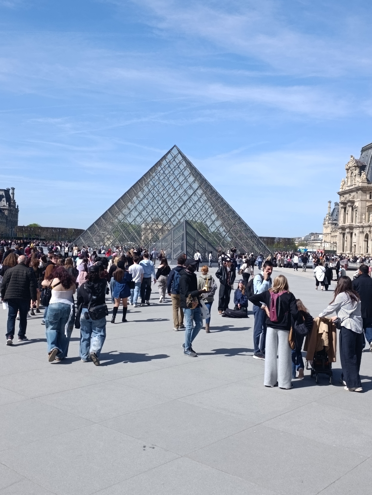 The image depicts a daytime scene at the Louvre Museum in Paris, France. In the background is the Louvre Pyramid, a large glass and metal structure. To the right is a portion of the Louvre Palace. The foreground is filled with a crowd of people walking and standing on a paved plaza. The sky is blue with sparse clouds. People are dressed in casual clothing. There is a stroller visible amidst the crowd.