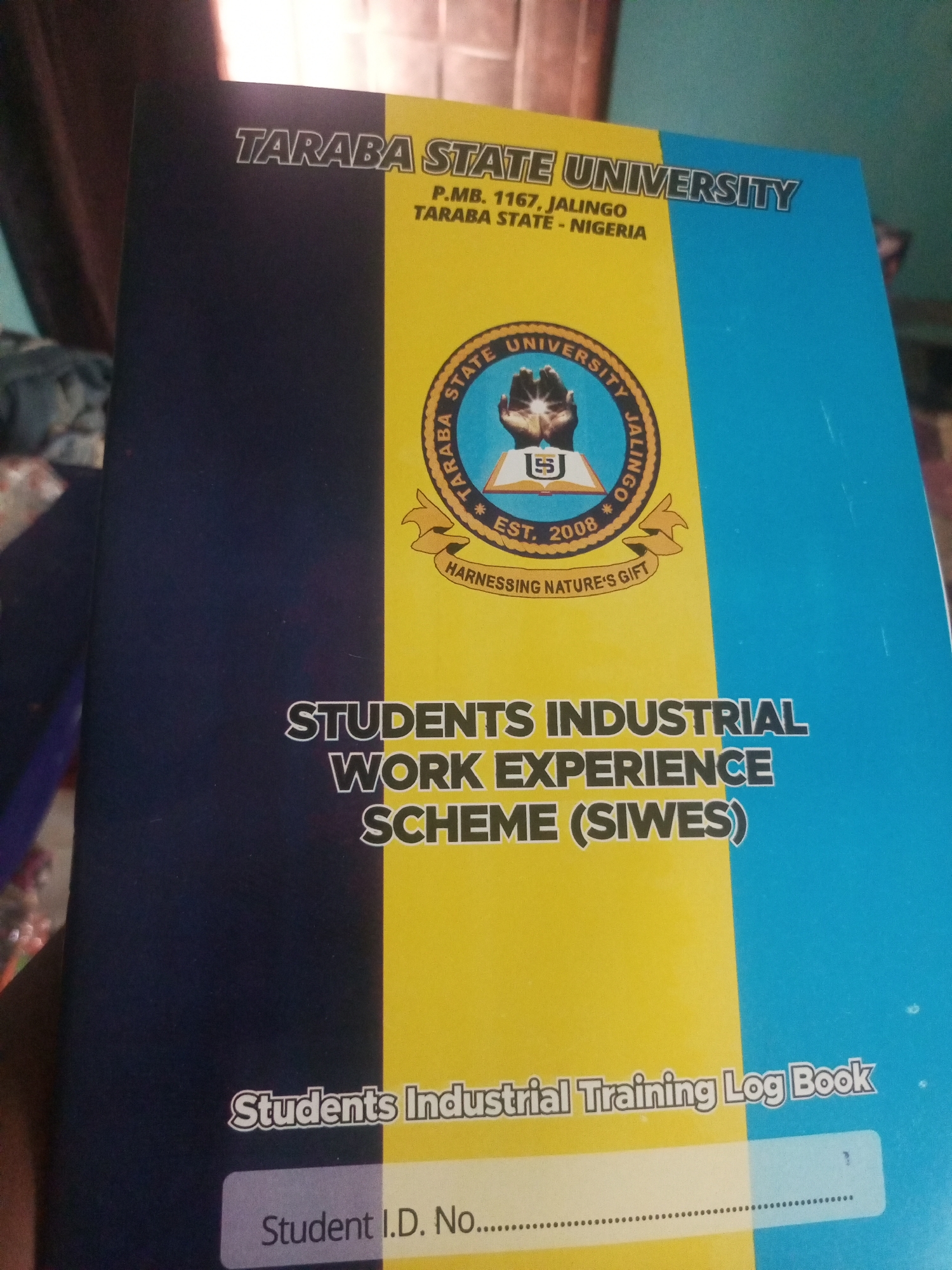 The image shows the cover of a "Students Industrial Training Log Book" from Taraba State University, located in Jalingo, Nigeria. The cover features the university's name prominently displayed at the top, followed by the address "P.M.B. 1167, Jalingo, Taraba State - Nigeria". Below this is the university's crest, which includes the text "Taraba State University Jalingo", "Est. 2008", and the motto "Harnessing Nature's Gift". The main title of the book is "STUDENTS INDUSTRIAL WORK EXPERIENCE SCHEME (SIWES)". At the bottom of the cover, it says "Students Industrial Training Log Book" and provides a space for "Student I.D. No....". The book cover is divided into two vertical sections: the left half is navy blue, and the right half is split between a bright yellow and a lighter blue.
