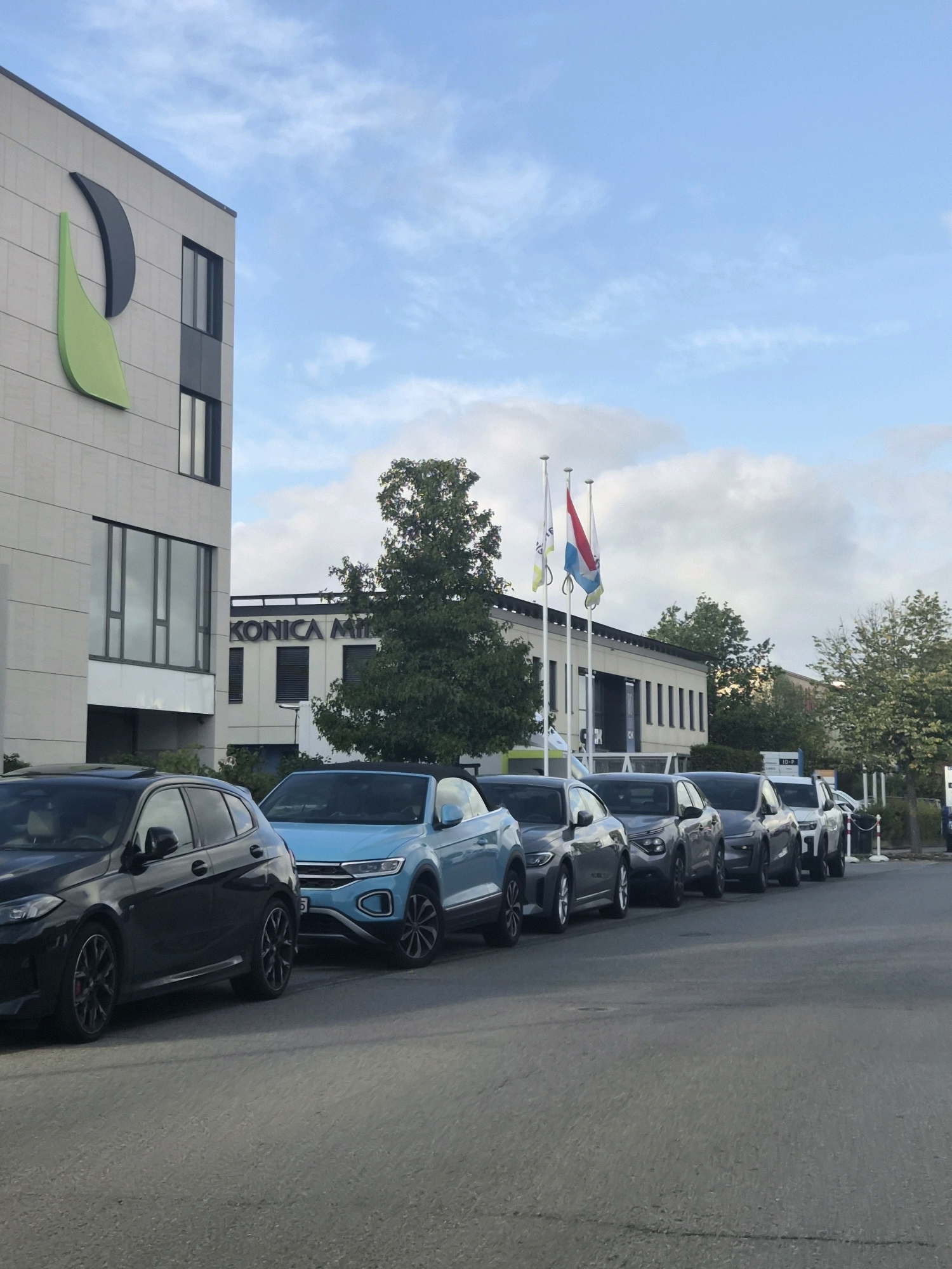 A line of cars is parked in front of a modern office building in Leudelange, Luxembourg. The building has a light-colored facade with large windows and a prominent green and gray logo on the left side. The name "KONICA MINOLTA" is visible on another section of the building. Three flags are planted in front of the building, one of which is the flag of Luxembourg. The sky is partly cloudy.