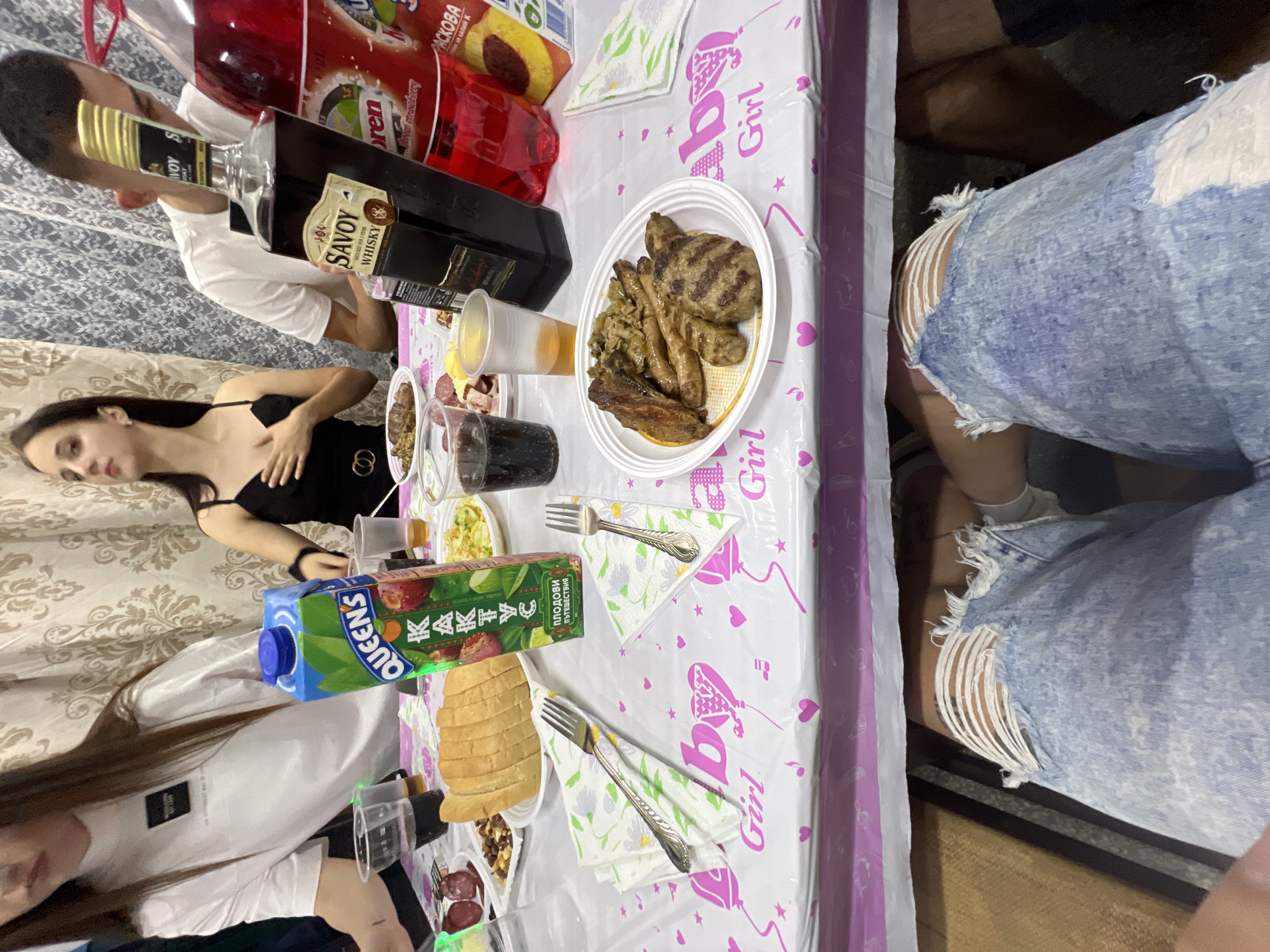 The image shows a table laden with food and drinks, suggesting a social gathering or meal. The tabletop has a white tablecloth with a repeating graphic pattern featuring "Girl" text and stylized imagery of hearts and musical notes.
Present on the table are multiple bottles of beverages, including what appears to be whiskey and various fruit juice boxes. A plate of grilled meats and sausages is prominently placed. There are also cups with liquid, plates of salad, bread, and other accompaniments like salami, indicative of a varied spread. Silverware is laid out on napkins, suggesting an organized setting for eating.
Two women are visible on the left side of the image, while a person holding a bottle is cropped at the top left. The jeans of another person with deliberate rips and exposed threads are visible on the top right. These elements suggest people are seated around the table, participating in the event.
There is no discernible information to suggest the image was taken specifically in the city of Trifonovo, Bulgaria.
