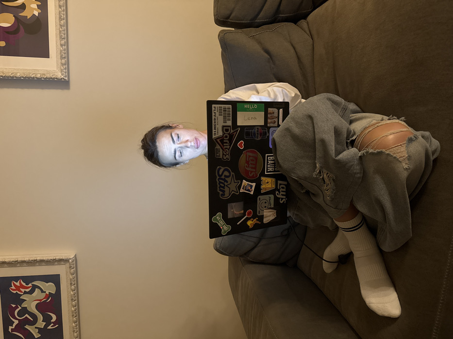An adult female is seated on a dark grey fabric sofa in an indoor setting in Venice, Italy. She wears a white long-sleeved top, light-wash ripped jeans with prominent tears on the knees, and white socks with blue stripes. Her hair is pulled back, and her face is illuminated by the light emanating from a black laptop positioned on her lap. Her gaze is directed downward towards the laptop screen. The laptop's lid is covered with numerous stickers, including discernible logos for "WE LOVE DOGS," "Star," "Lays," "Doritos," "Pepsi," and a sticker displaying "PLPF4H3M1G" and the name "Lena." A charging cable extends from the laptop. The background consists of a plain, light-colored wall. Two framed abstract art pieces are mounted on the wall; one on the upper left features red, white, and purple shapes, and another on the upper right displays purple and yellow organic forms. The room is dimly lit, with the primary light source being the laptop screen.