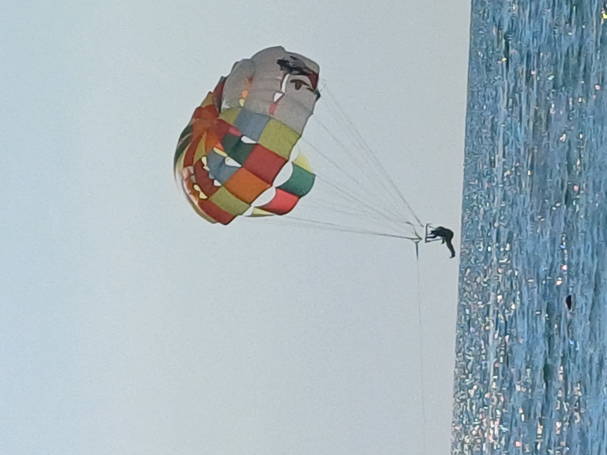 A person is parasailing high in the sky. They are attached to a colorful parachute, which is adorned with a patchwork of red, orange, yellow, green, and blue squares, as well as some grey sections that appear to depict a face. The parachute is being pulled by unseen forces, likely a boat, as indicated by the taut lines extending from the parachute harness to the right of the frame. The person is wearing dark clothing and is suspended in the air, facing away from the camera.
To the right of the parachute and person, a large, vertical surface dominates the right side of the image. This surface is textured and appears to be made of numerous vertical, shimmering strands, possibly a decorative curtain or a building facade with a reflective, iridescent quality. The color is predominantly blue with highlights of white and pink, suggesting it is catching the light.
The sky is a pale, washed-out blue, indicating a bright day, possibly late morning or afternoon given the light. The overall scene suggests an outdoor recreational activity taking place at a location that offers such attractions. The presence of the shimmering blue surface on the right could be a clue to the location, perhaps a waterfront resort or a venue with elaborate decorations. There is no visible text in the image. The activity is one of leisure and adventure, with the person seemingly enjoying the experience of flying high above the ground.
