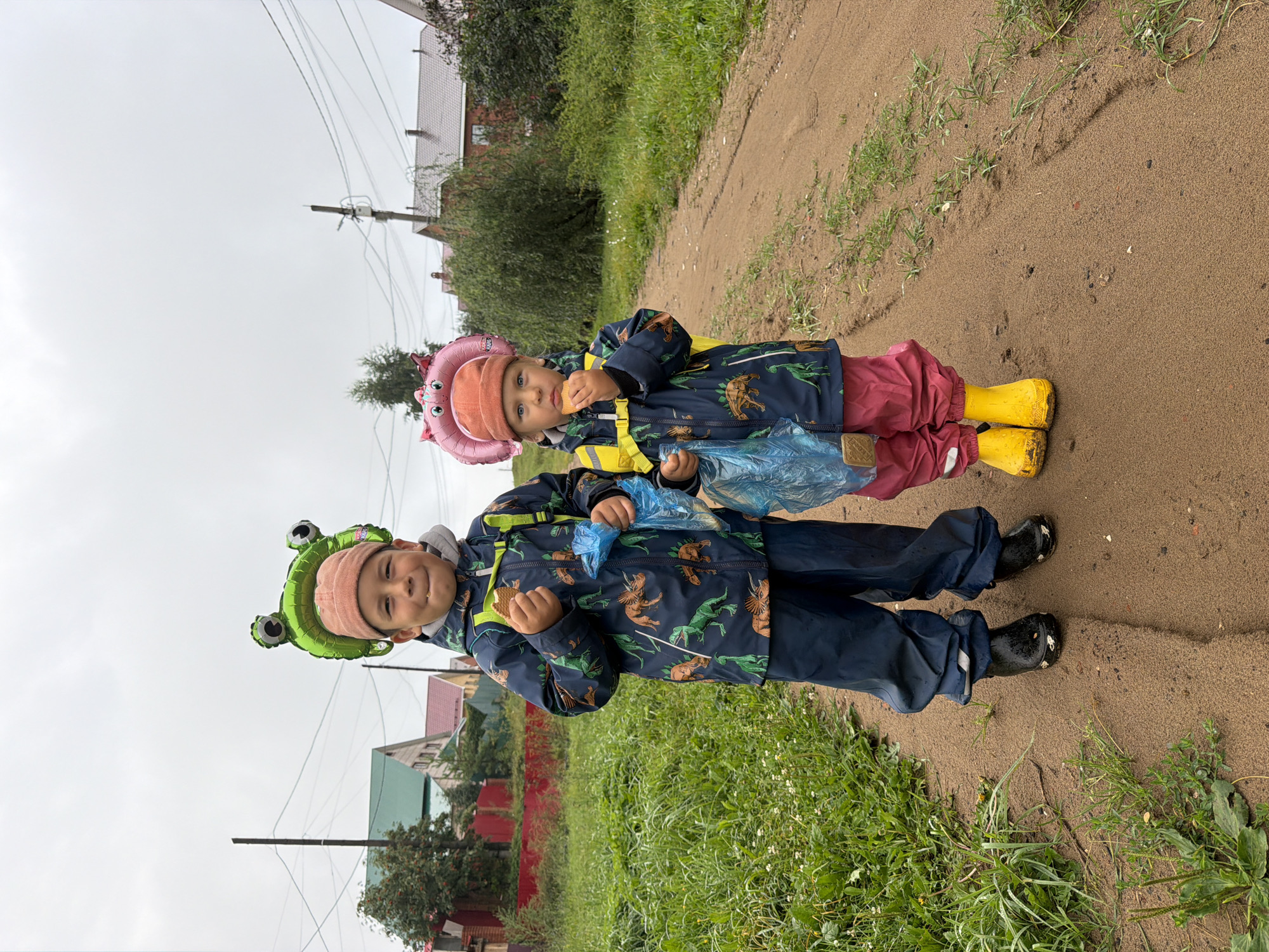 Two children, a boy and a girl, stand on a dirt path outdoors under a cloudy sky. The boy, on the left, wears a dinosaur-print jacket, dark blue rain pants, and black rain boots. He has a green frog-shaped balloon above his head and is holding a cookie and a blue plastic bag. The girl, on the right, wears a pink balloon headpiece, a jacket with a similar dinosaur print, pink rain pants, and yellow rain boots. She is also holding a cookie and a blue plastic bag. The background shows houses and trees, indicative of a rural or suburban setting. The location is identified as the city of Chaikovsky, Russia.
