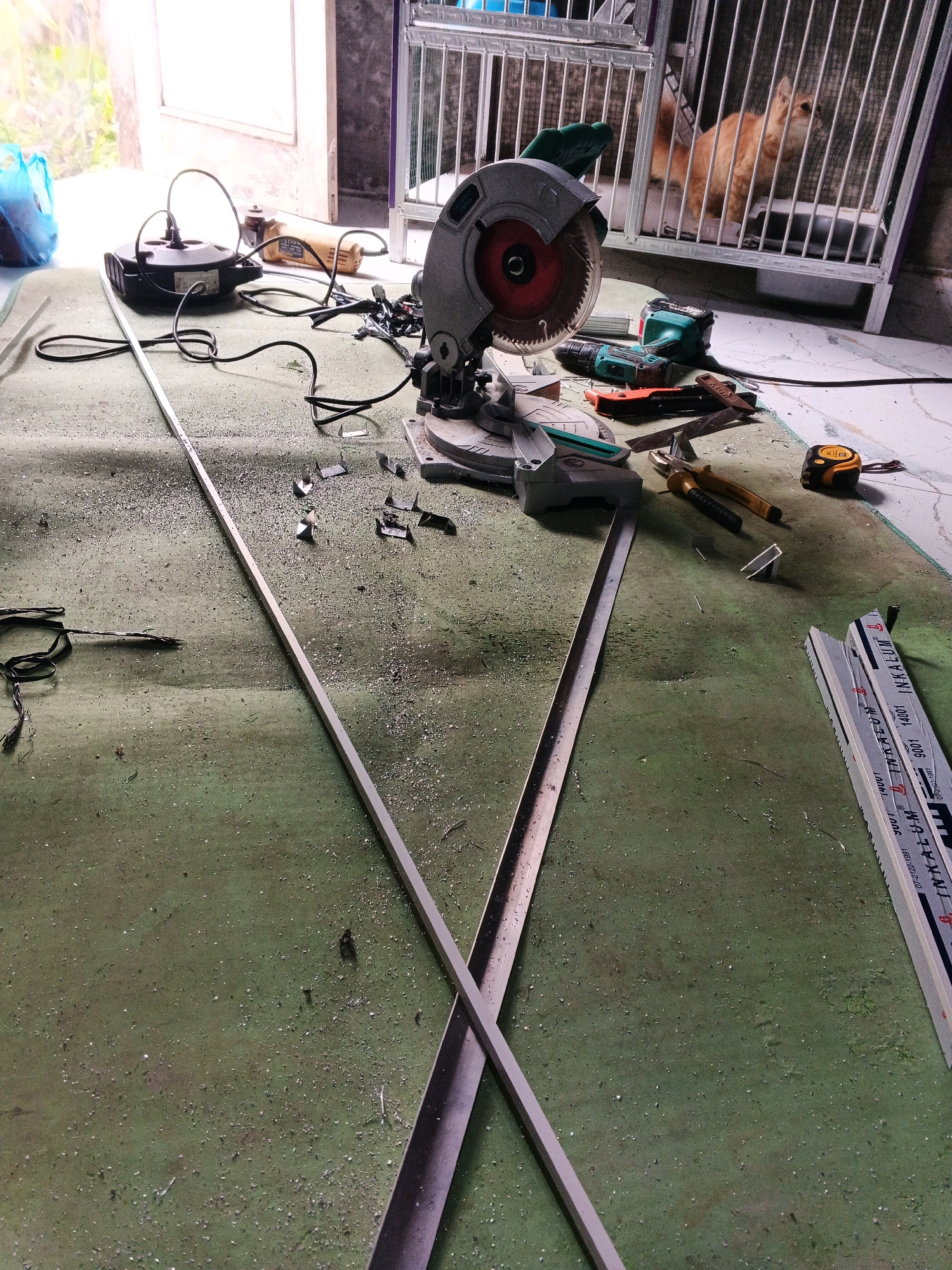In an indoor construction or workshop area, possibly in Planjan, Indonesia, a variety of tools and materials are scattered across a green, somewhat dirty floor, covered in metal shavings. A large compound miter saw, a power drill, pliers, a tape measure, and an angle grinder are present, suggesting recent cutting or assembly work. Two long, thin metal pieces are laid out on the floor, crossed over each other, with one piece extending towards the viewer. In the background, an orange cat is visible inside a white metal cage, and an open doorway leads to a brightly lit outdoor area. A blue plastic bag is also visible near the doorway. The overall impression is one of an active work area.