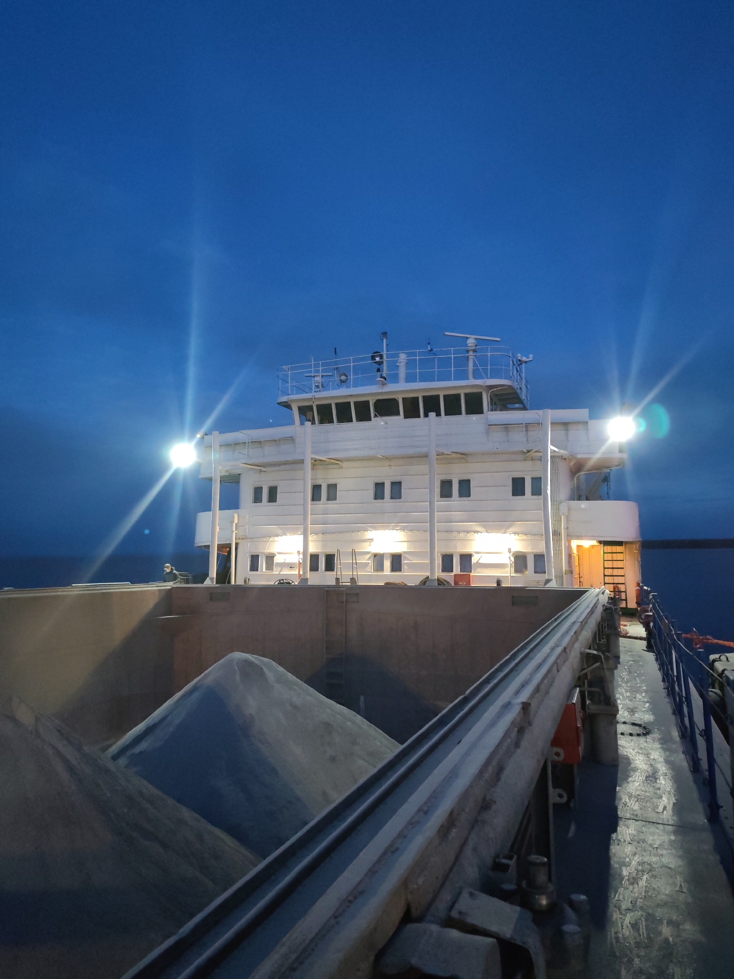 The image depicts the deck of a barge, possibly near Shcheleiki, Russia. A large white superstructure rises from the deck, illuminated by several lights, creating bright glare points in the image. The barge is loaded with what appears to be a gray granular material, possibly gravel or sand, forming a mound in the center. A mechanical conveyor belt runs along the deck. The sky is a deep blue, indicating either twilight or a night scene. A body of water is visible at the edge of the deck.