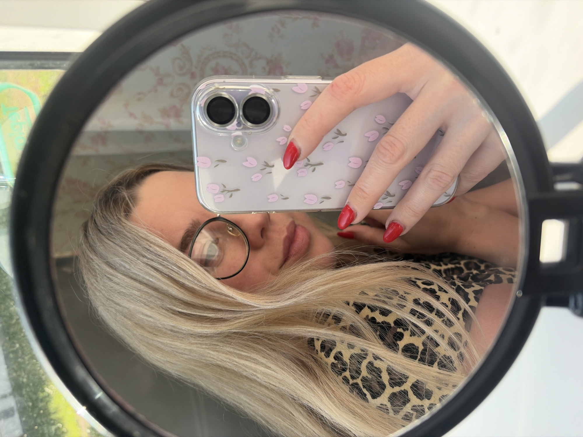 A woman with long blonde hair and red nail polish is taking a selfie in a round mirror. She's wearing a black-rimmed eyeglass on her left eye and a leopard print top. She is holding a phone with a clear case adorned with small pink flowers. The reflection in the mirror also shows a blurred background with a window showing greenery. The setting appears to be indoors. There is no indication of Chaikovsky, Russia.