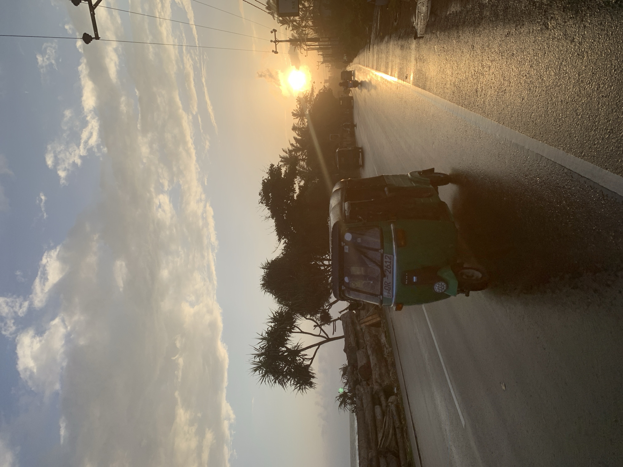 The image depicts a street scene in Ahangama, Sri Lanka, during what appears to be sunset. A green auto-rickshaw, identified by the license plate "SP-QR-2612", is prominently featured in the foreground, driving on a wet asphalt road. The road reflects the golden light of the setting sun, creating a shimmering effect. In the background, palm trees and other tropical vegetation line the street. A line of utility poles with wires stretches along the right side of the road. The sky is partly cloudy with the sun visible near the horizon, casting a warm glow over the scene. To the left, a rough embankment of logs and driftwood is visible near the ocean. Other vehicles, including motorcycles and cars, are present further down the road, indicating moderate traffic. The overall atmosphere is serene and evocative of a tropical evening.